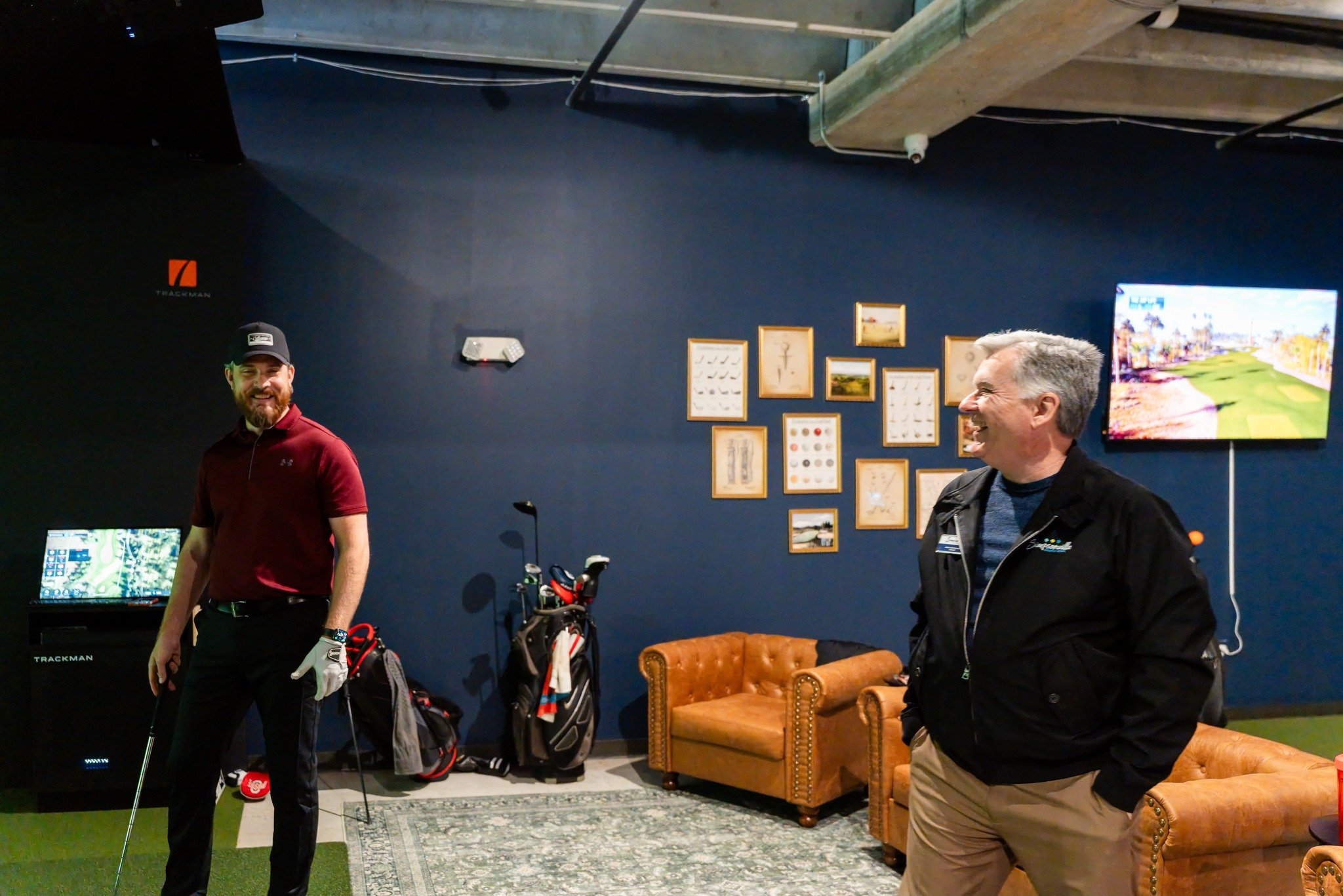 Two men in a golf-themed indoor setting, one wearing a red shirt and cap, the other in a black jacket, smiling and standing near golf clubs, framed pictures, and a television displaying a golf course.