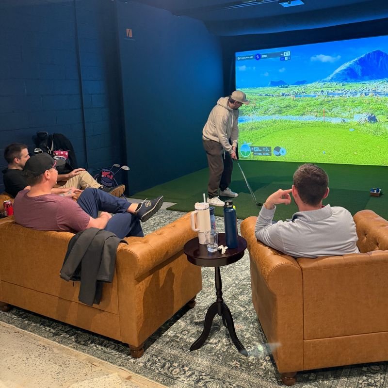 People playing indoor golf simulation game with a large screen showcasing a virtual golf course, seated on brown couches in a lounge area.