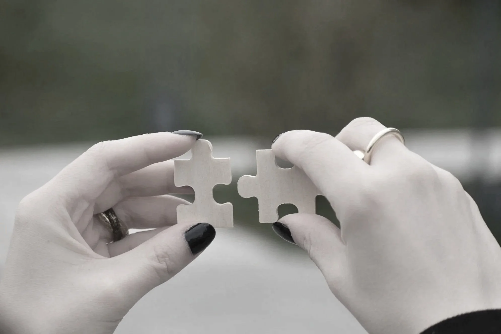 Two hands with black painted nails hold puzzle pieces, about to connect them, with a blurred outdoor background.