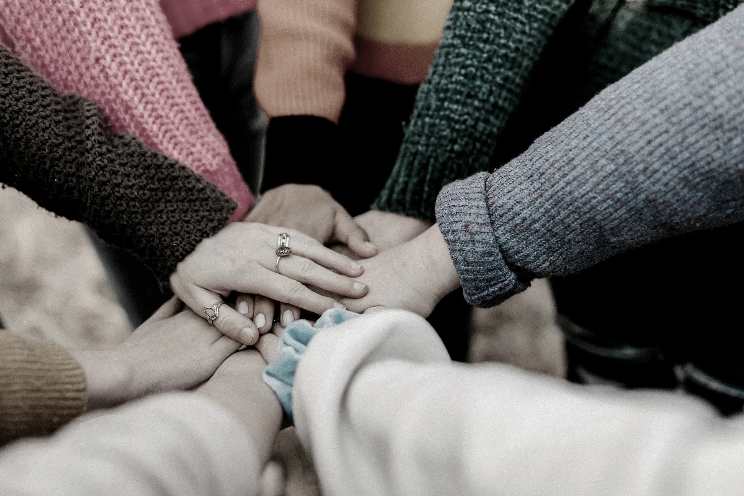 A close-up of multiple people holding hands, forming a circle, including a baby with a hospital bracelet, wearing various long-sleeve shirts, symbolizing support and connection.
