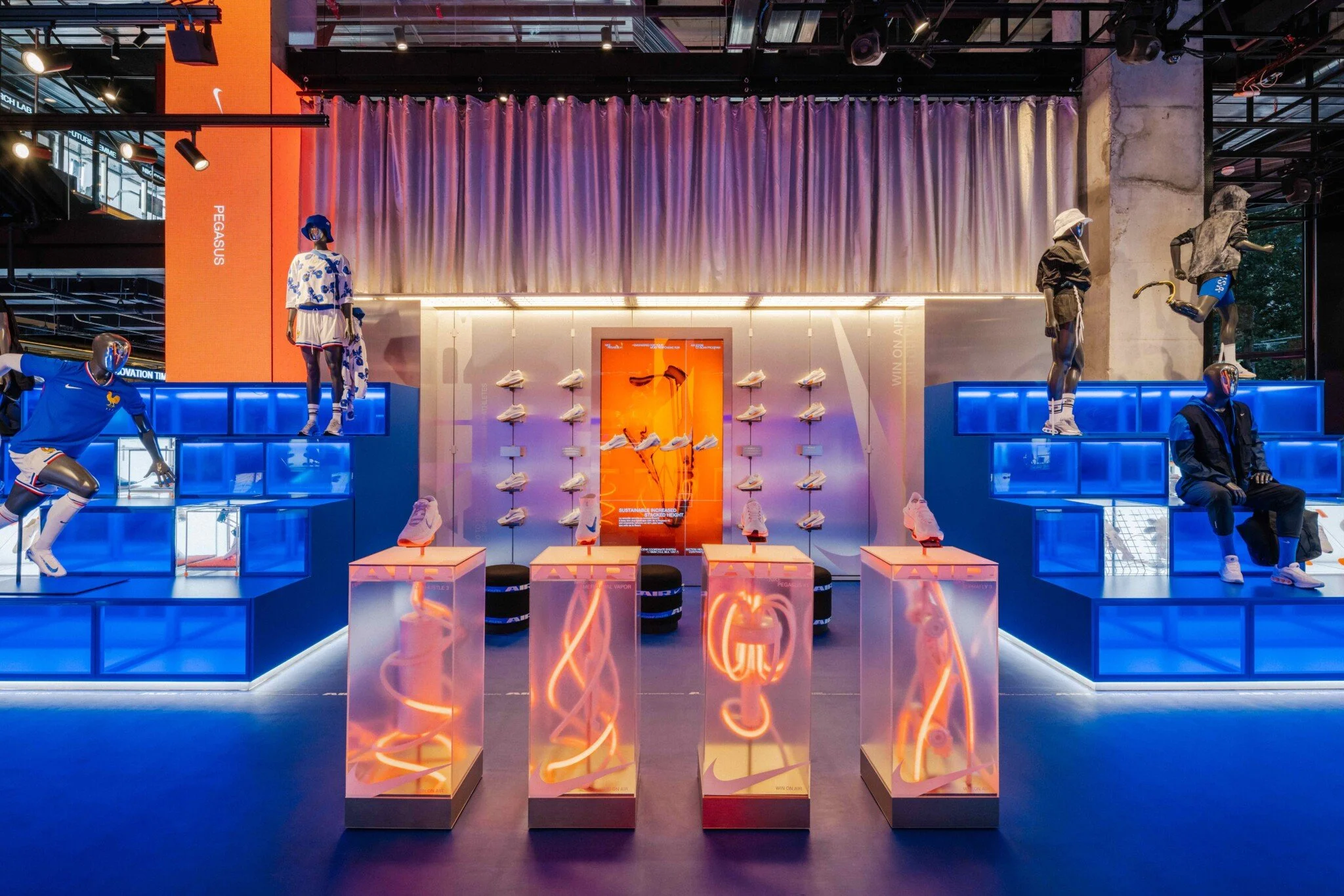An indoor retail display featuring mannequins dressed in athletic clothing, with a prominently lit Nike section, shoes on shelves, and neon signs, all illuminated with blue and purple lighting.