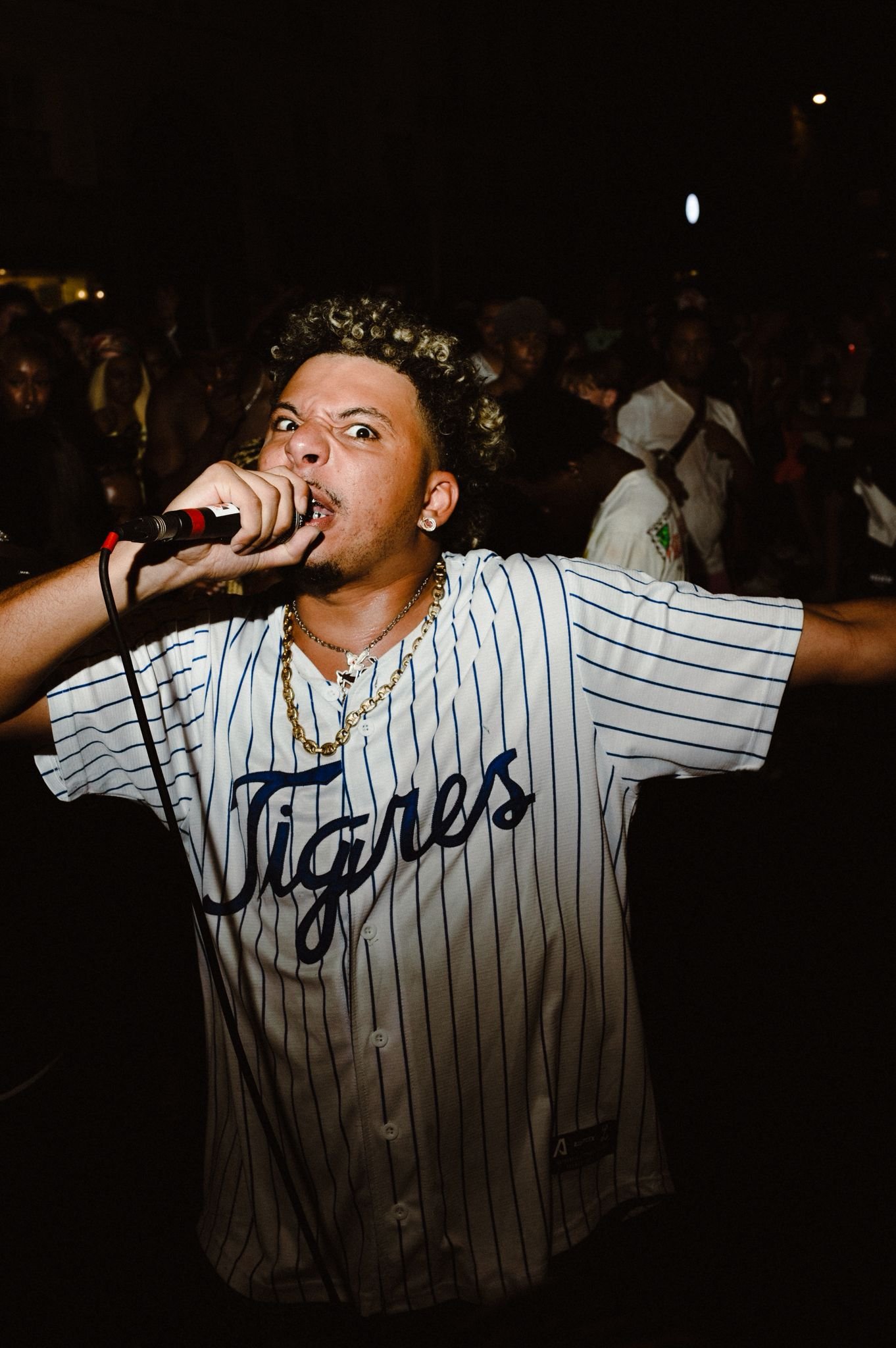 A man with curly hair, tattoos, and gold jewelry, wearing a baseball jersey, holding a microphone, and singing passionately at a nighttime event with a crowd in the background.