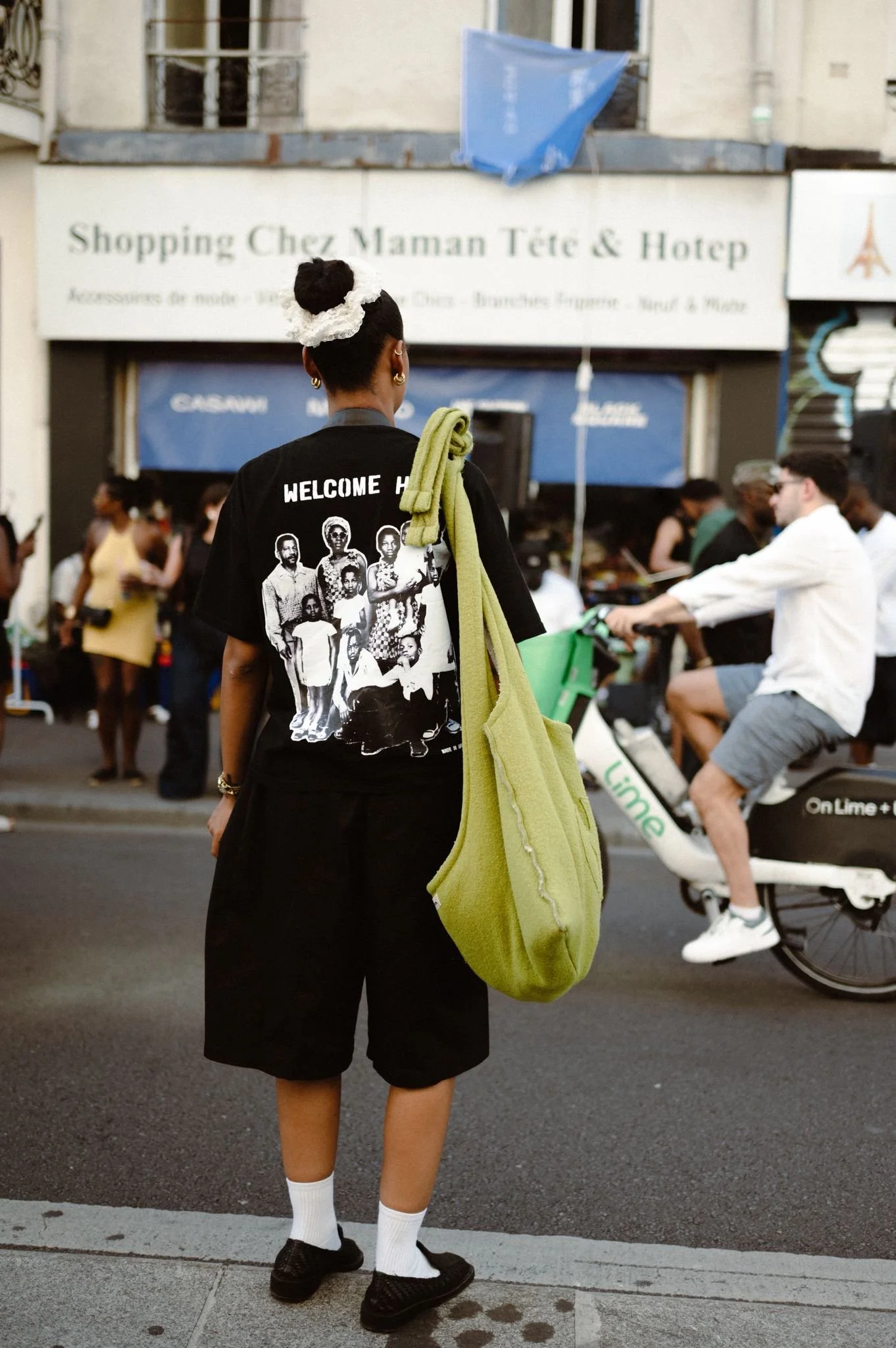 A girl with a bun hairstyle decorated with lace, wearing a black T-shirt with graphics and the words 'WELCOME HOME,' black shorts, white socks, and black sneakers, standing on a city sidewalk with people around and blurred storefronts in the background.