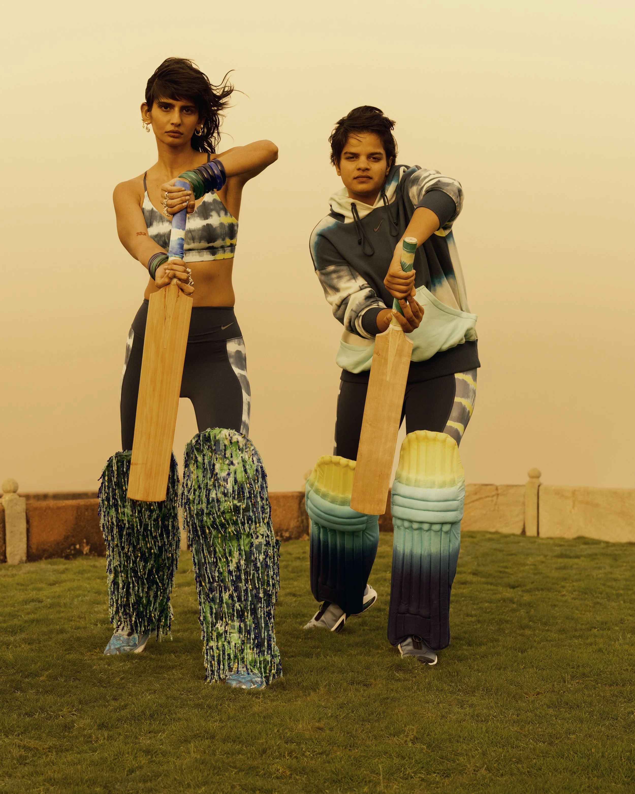 A woman and a young man are in a field, holding wooden sticks. The woman has short, wavy hair, multiple bracelets, and is wearing a patterned crop top and patterned pants. The young man has short, dark hair, and is wearing a hoodie with a design and loose pants with a gradient pattern. They are standing on grass with a plain sky background.