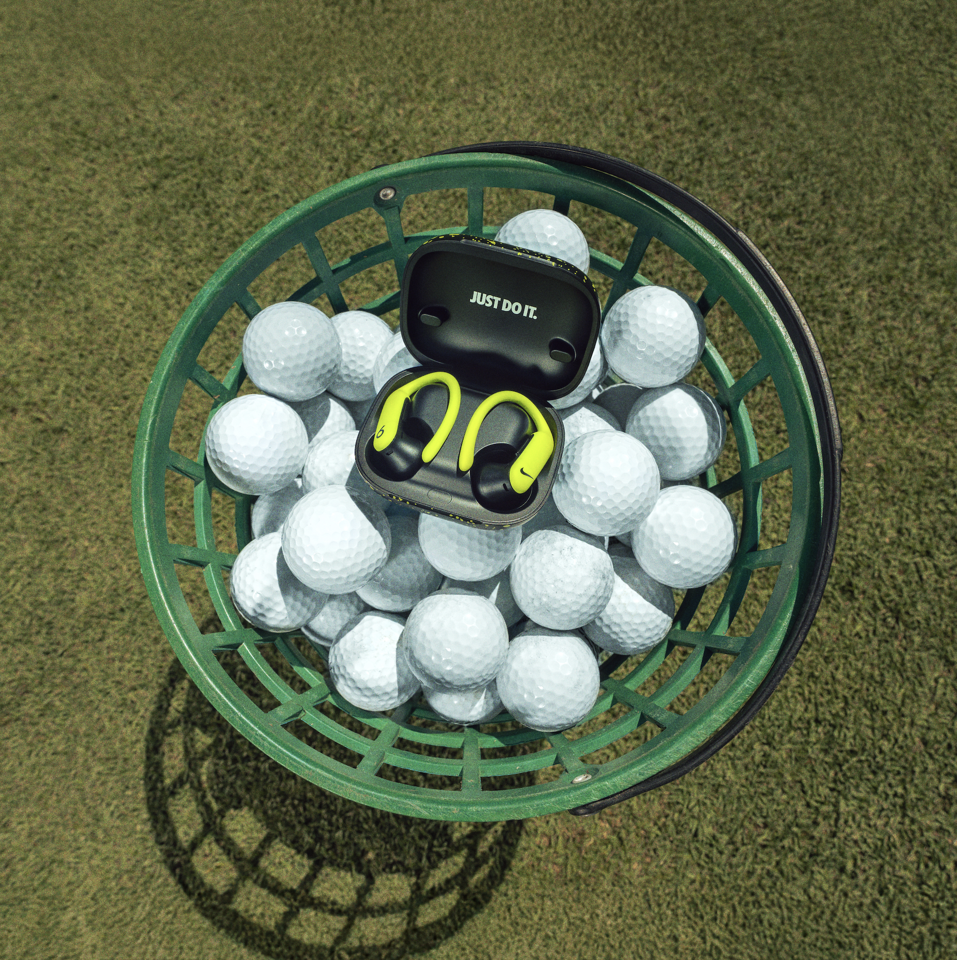Green basket filled with white golf balls and a black case of yellow and black wireless earbuds, with a grassy surface underneath.