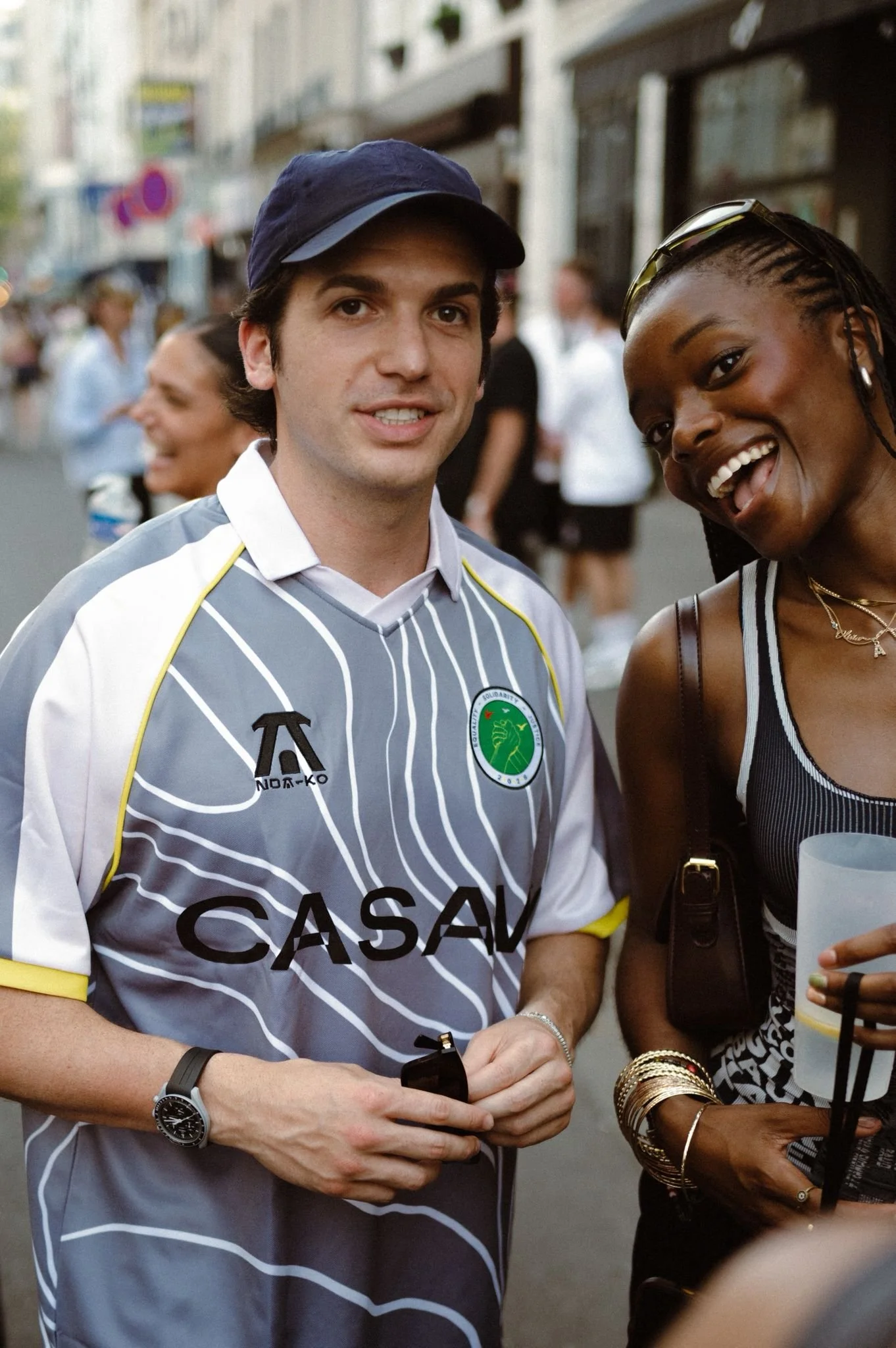 Two people standing and smiling on a city street. The man is wearing a gray and white striped soccer jersey with a green and white badge, and the woman is wearing a black and white striped outfit with gold jewelry.