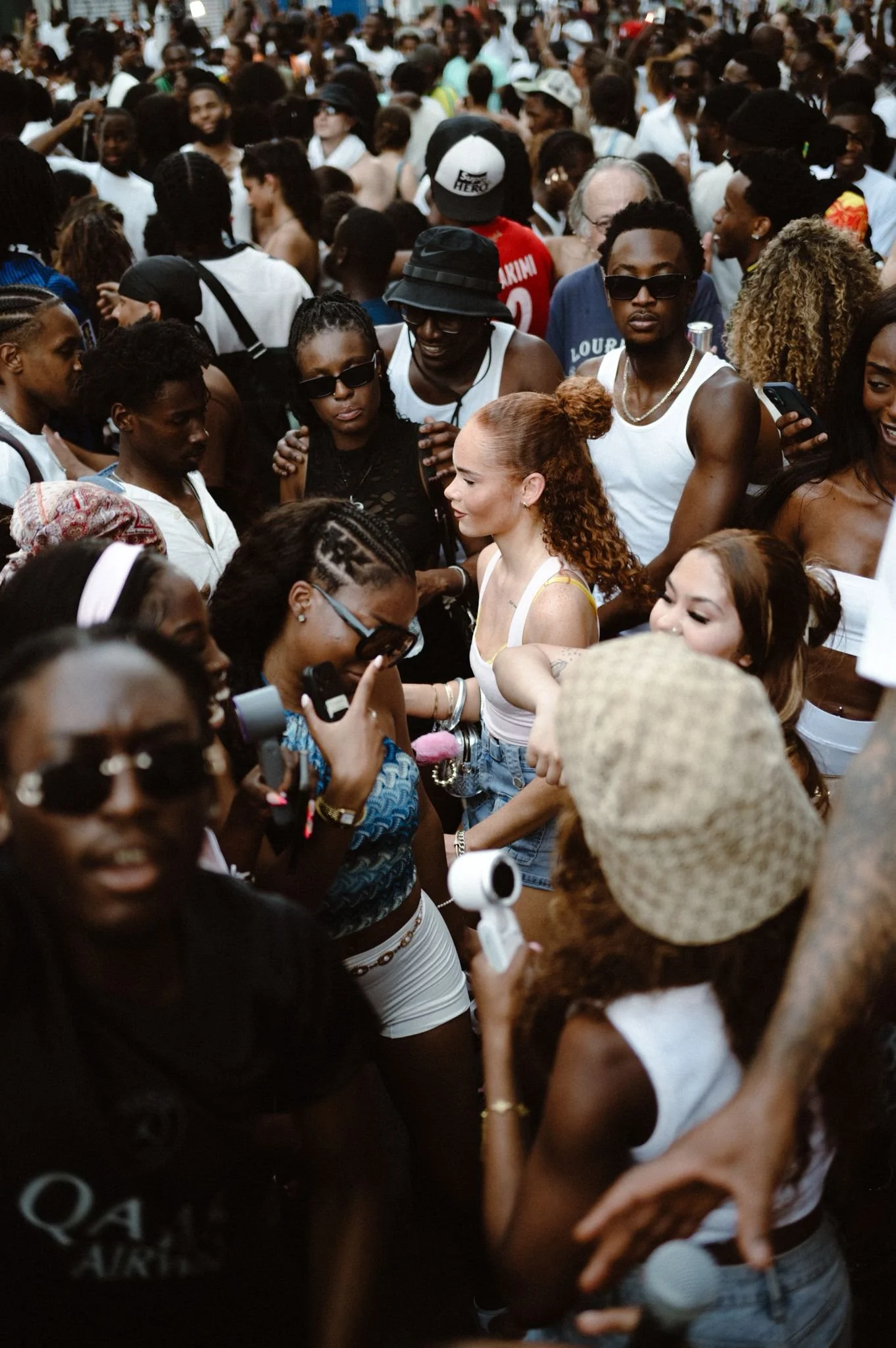 Crowd of diverse people gathered outdoors, some taking pictures and engaging with each other, wearing casual summer clothing.
