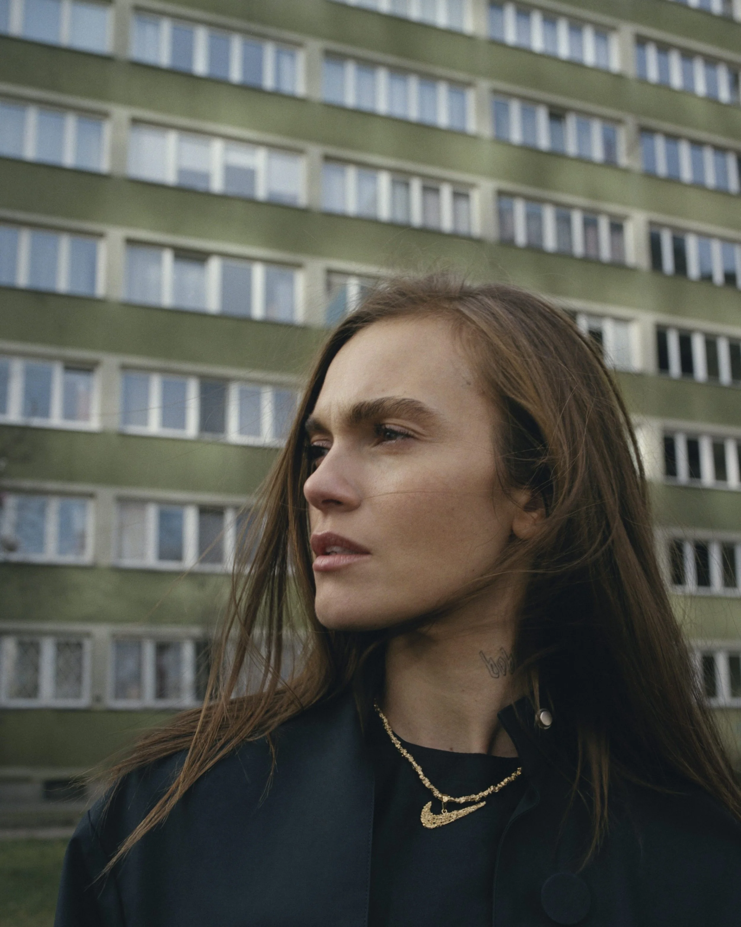 Close-up of a young woman with long brown hair, wearing layered necklaces and a black jacket, standing outdoors in front of a green apartment building with white balconies.