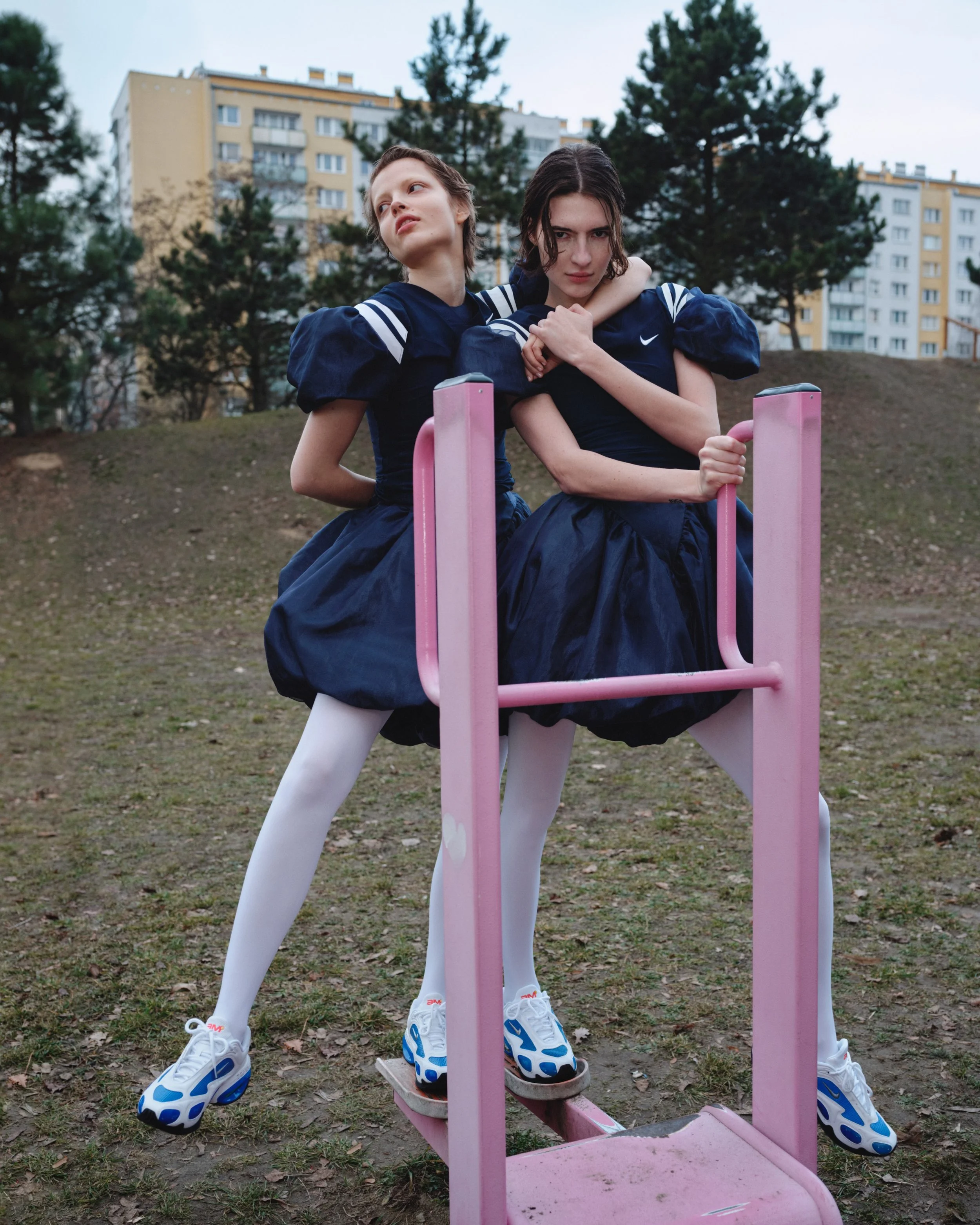 Two young women with short brown hair wearing navy blue dresses with white stripes on the sleeves, white tights, and sneakers standing on a pink playground see-saw in an outdoor park with trees and residential buildings in the background. One woman is holding the handle of the see-saw while the other stands behind her with her arm around her shoulders.