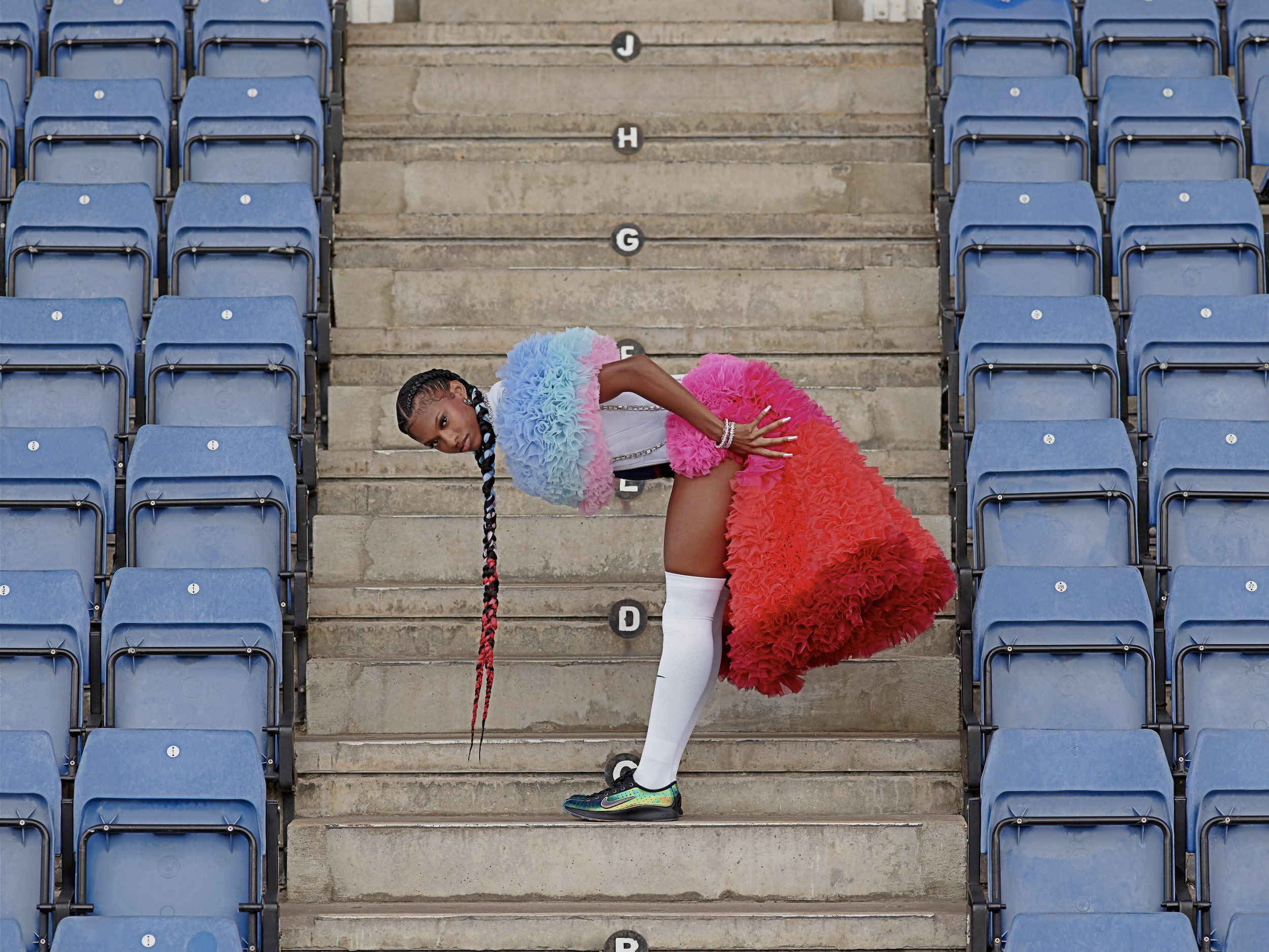 A young woman in athletic clothing and colorful sneakers posing on stadium stairs with blue seats on either side, wearing a large rainbow-colored ruffled skirt, white tights, and a pastel ruffled top, leaning forward with one hand on her hip and the other on her thigh.