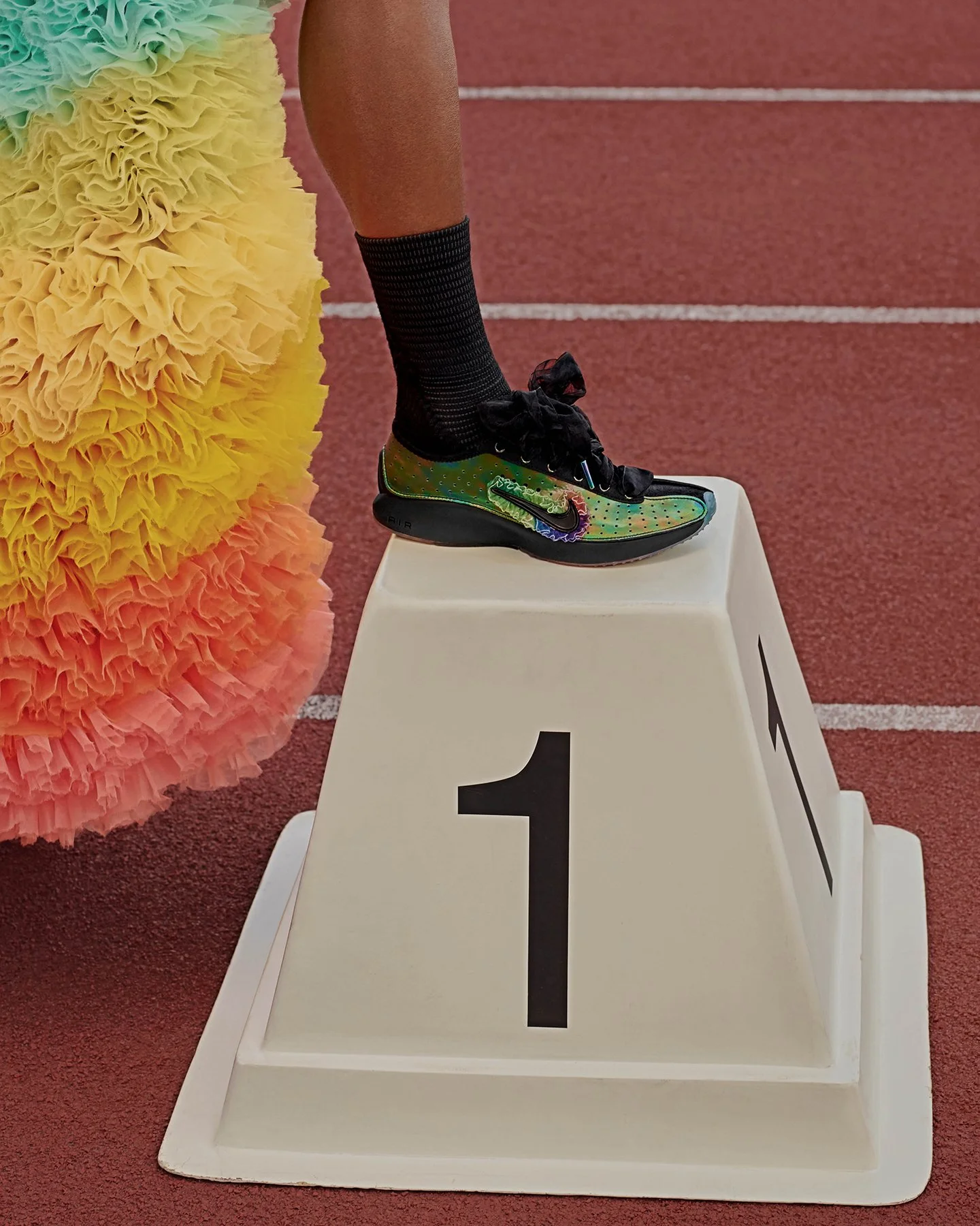 Close-up of a runner's foot on the first-place podium at an athletic event, with a colorful, iridescent running shoe, black socks, and part of a yellow, orange, pink, or peach pom-pom decoration nearby on a red track.