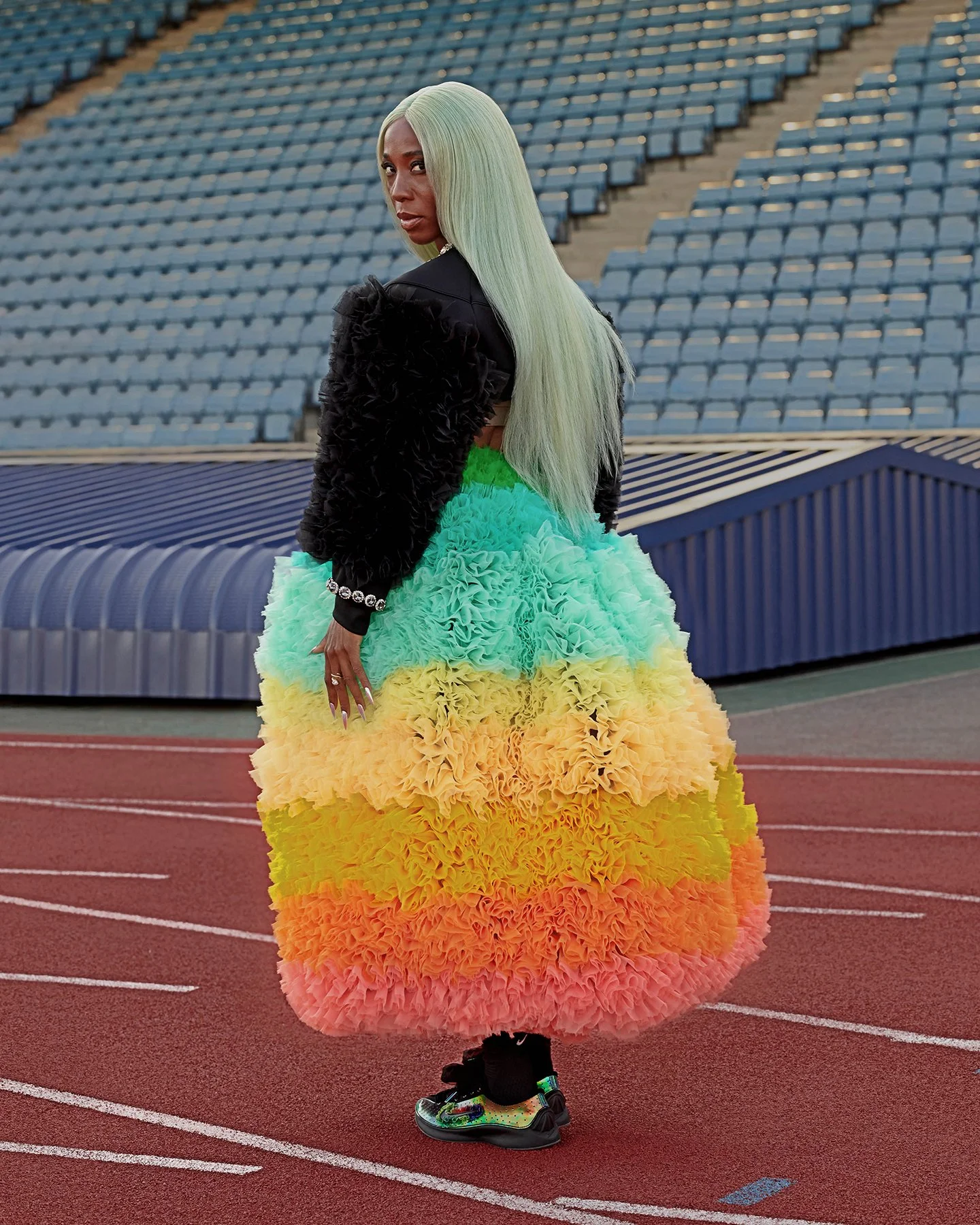Woman standing on a running track wearing a colorful, fluffy, tiered gown with rainbow colors, black jacket, and sneakers.