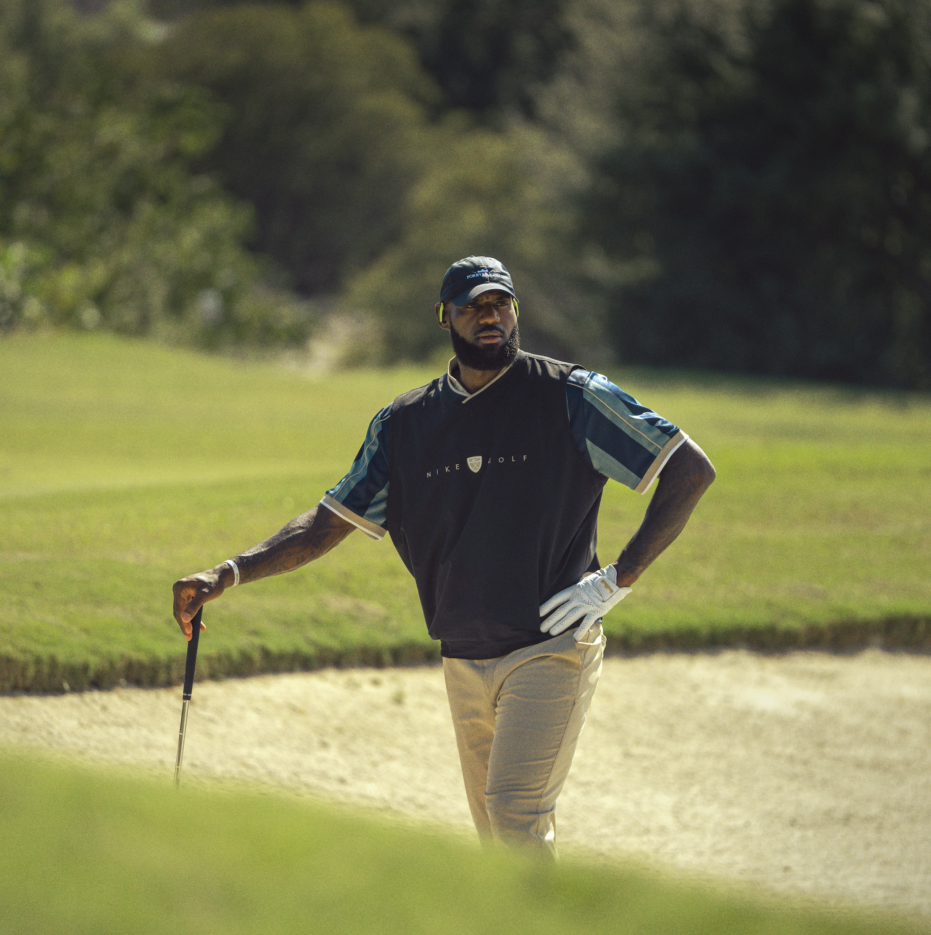 A man playing golf on a golf course, standing on a sand trap, wearing a Nike Golf shirt, cap, and headphones, holding a golf club.