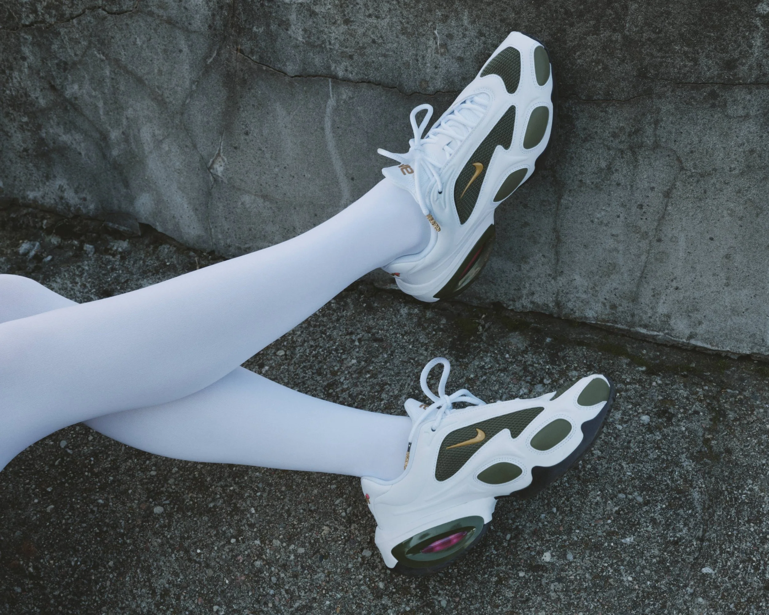A person standing outdoors on rocky terrain, wearing white athletic shoes with olive green and gold accents, and white leggings.