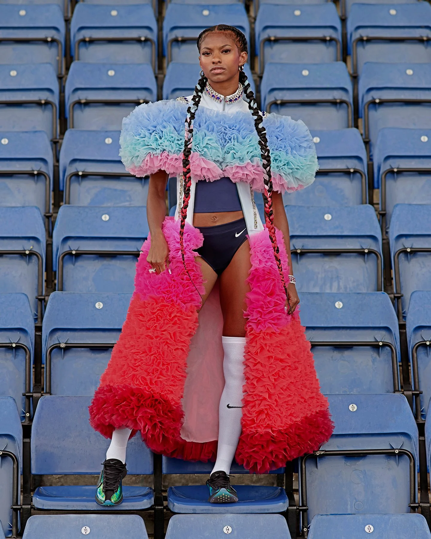 A woman standing alone in an empty stadium seating area, wearing a colorful, ruffled outfit with a long, bright red and pink coat, Nike socks, and sneakers.