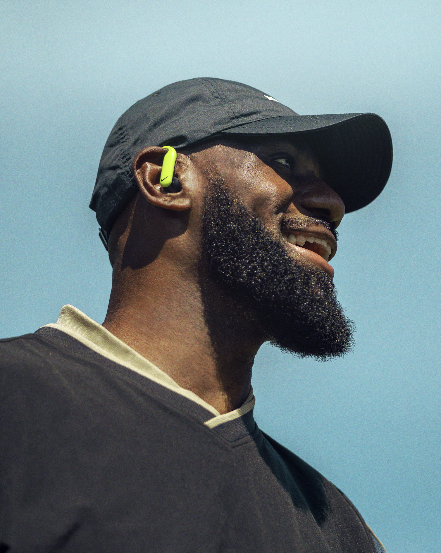 Close-up of a man wearing a black cap and a gray shirt with a beige collar, smiling, with a neon yellow Nike wireless earbud in his ear, against a clear blue sky.