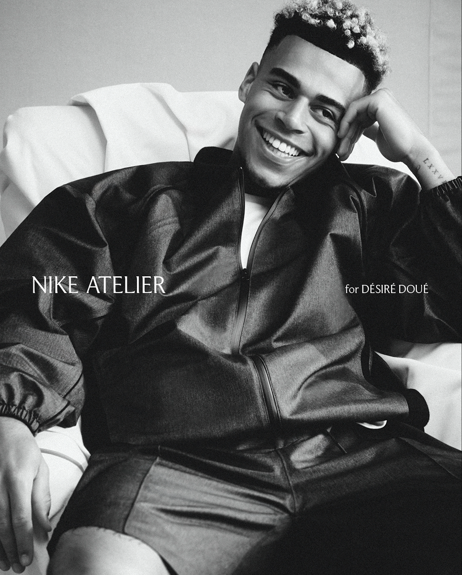 A smiling young man with short hair, wearing a leather jacket, sitting in a room with a light-colored wall. The photo is in black and white.