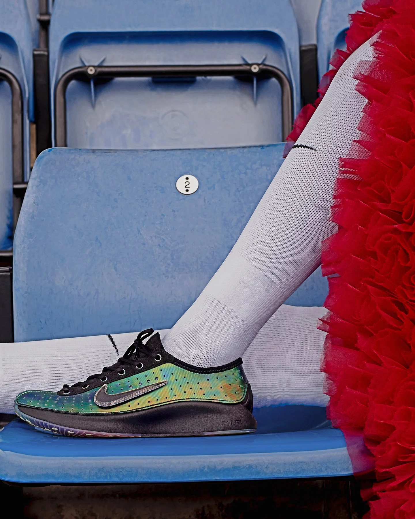 An ice skating rink seat with a blue plastic chair, part of a figure skater's leg in a white tights and colorful skate shoe, and a red feather boa on the right side.