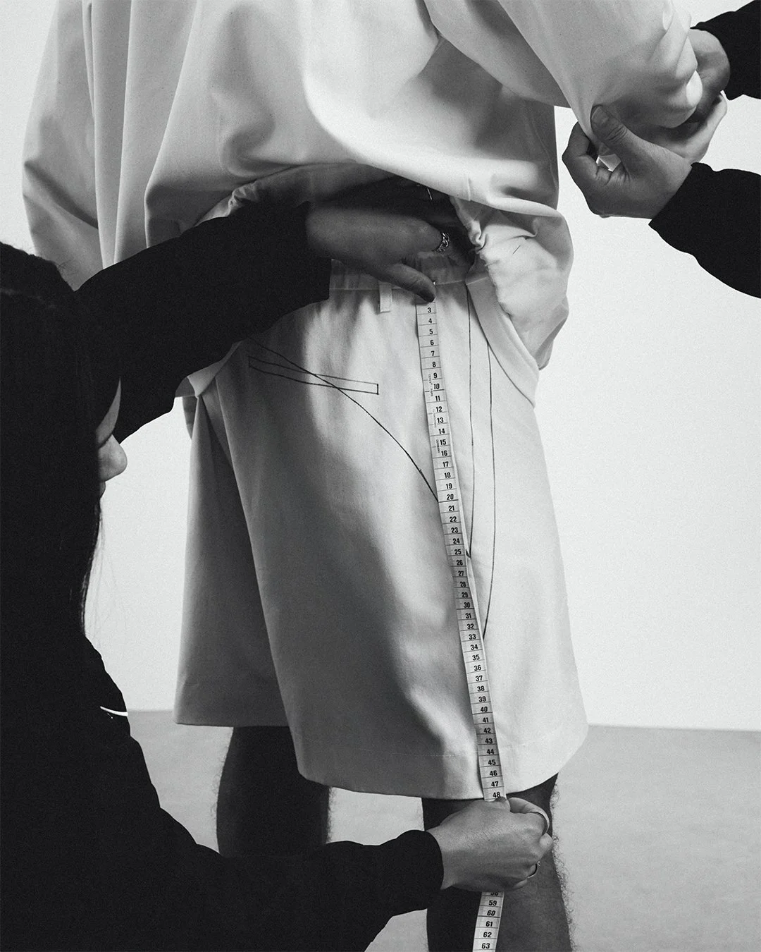 Black and white photograph showing three people measuring a man's waist and leg with a measuring tape. The man is wearing a white shirt and white shorts, and the others are dressed in dark clothing, one with a hood.