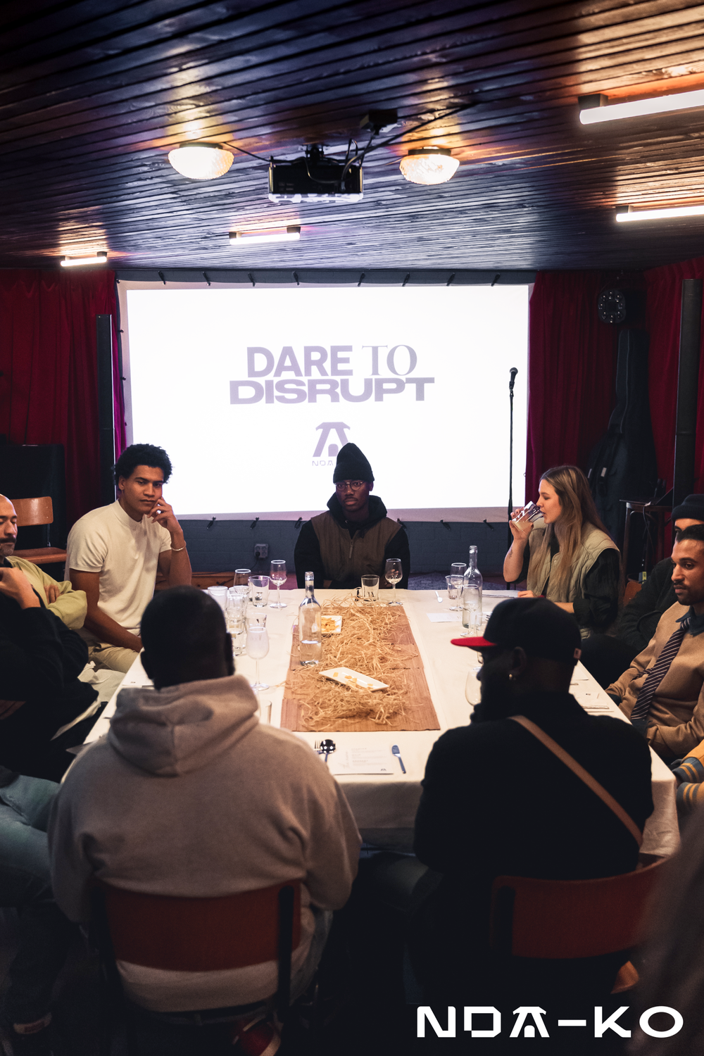 People gathered around a conference table during a meeting or panel discussion, with a large screen in the background displaying the text 'DARE TO DISRUPT' and a logo.