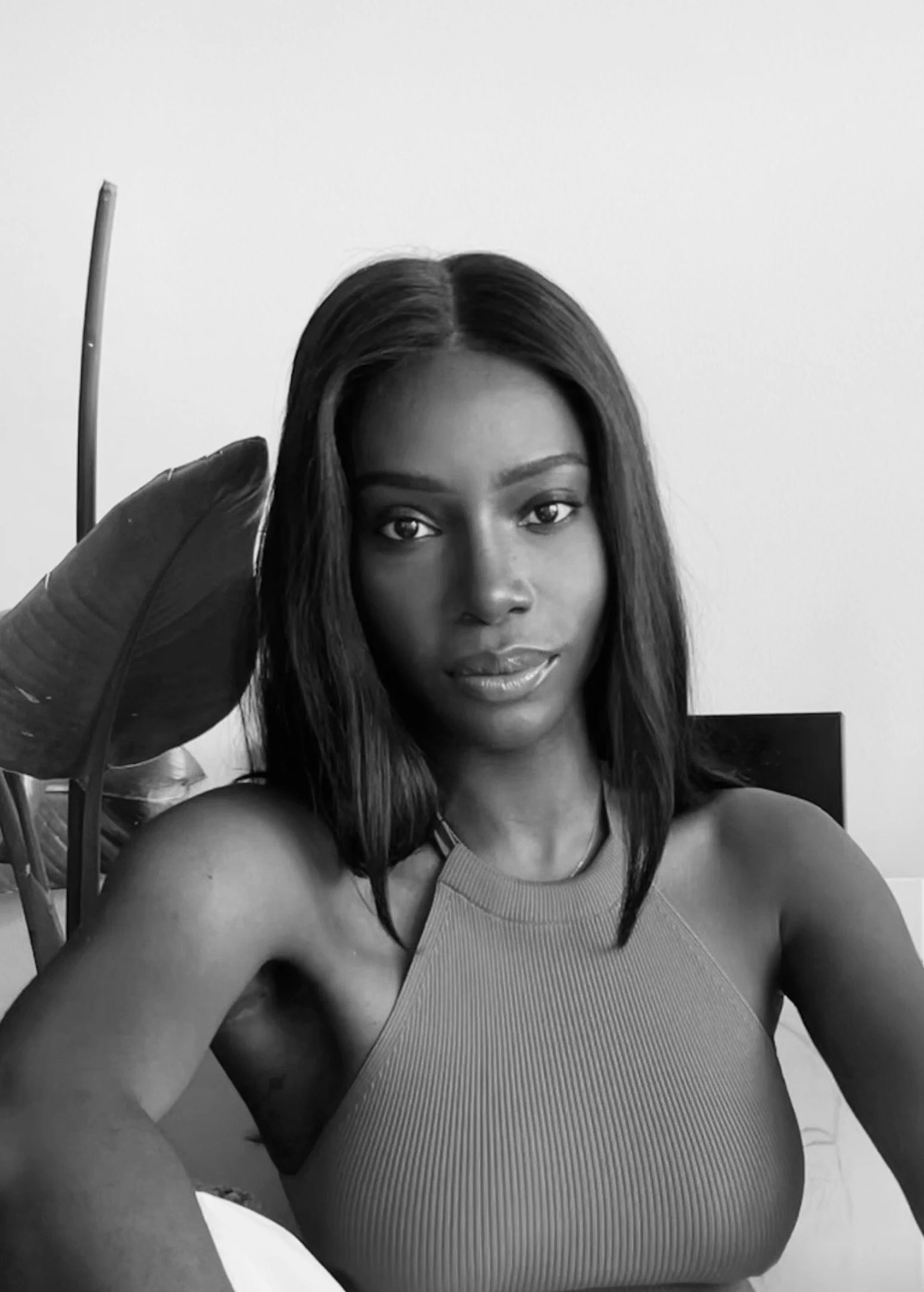 Black and white photo of a woman taking a selfie, wearing a sleeveless top, with a large leaf partially covering her head and shoulder, and a plain background.
