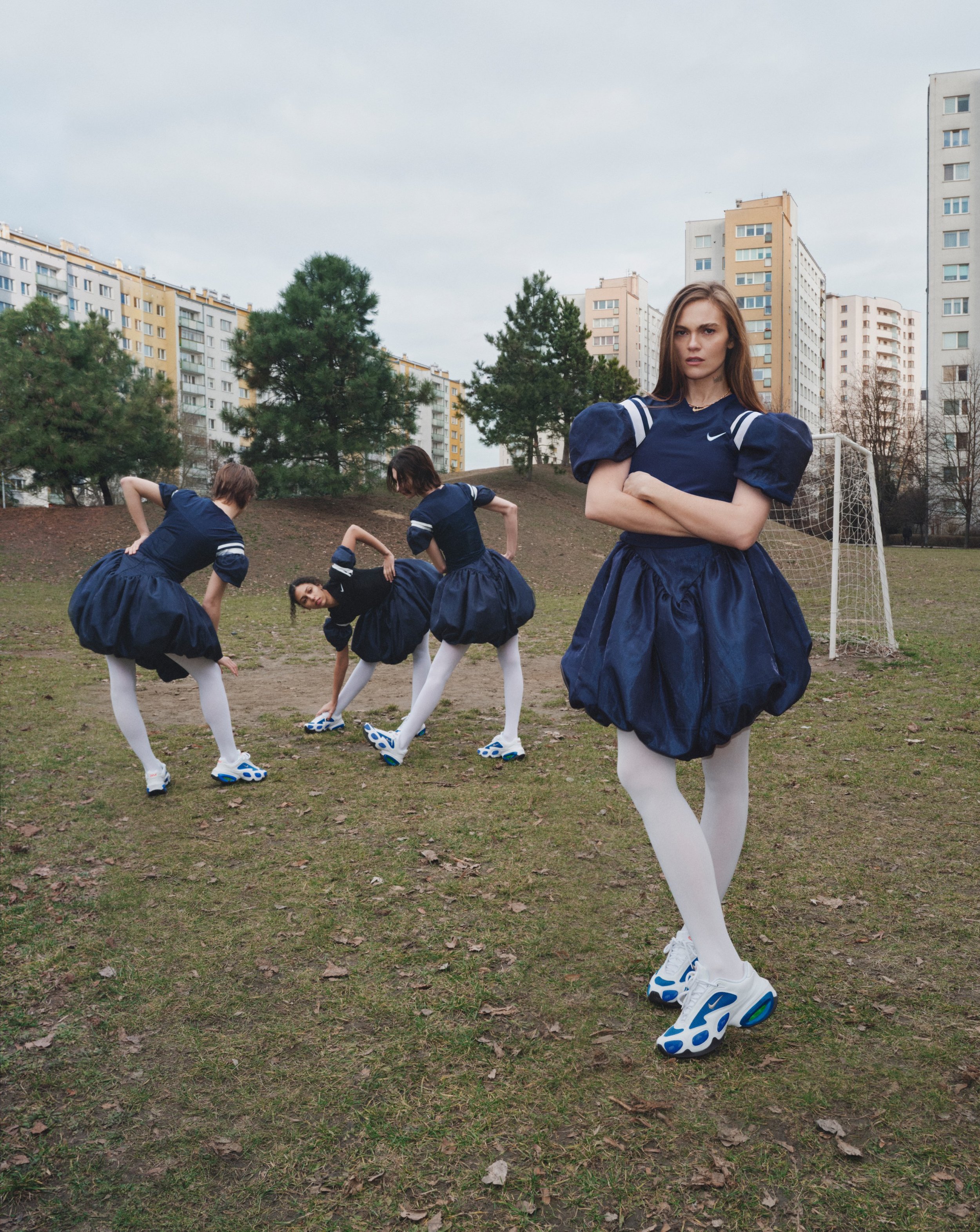 Four women in matching navy blue sports uniforms and white leggings, wearing white sneakers with blue accents, are on a grassy field with trees and tall apartment buildings in the background. Three women are bending over touching their toes, while one woman stands with arms crossed, looking at the camera.