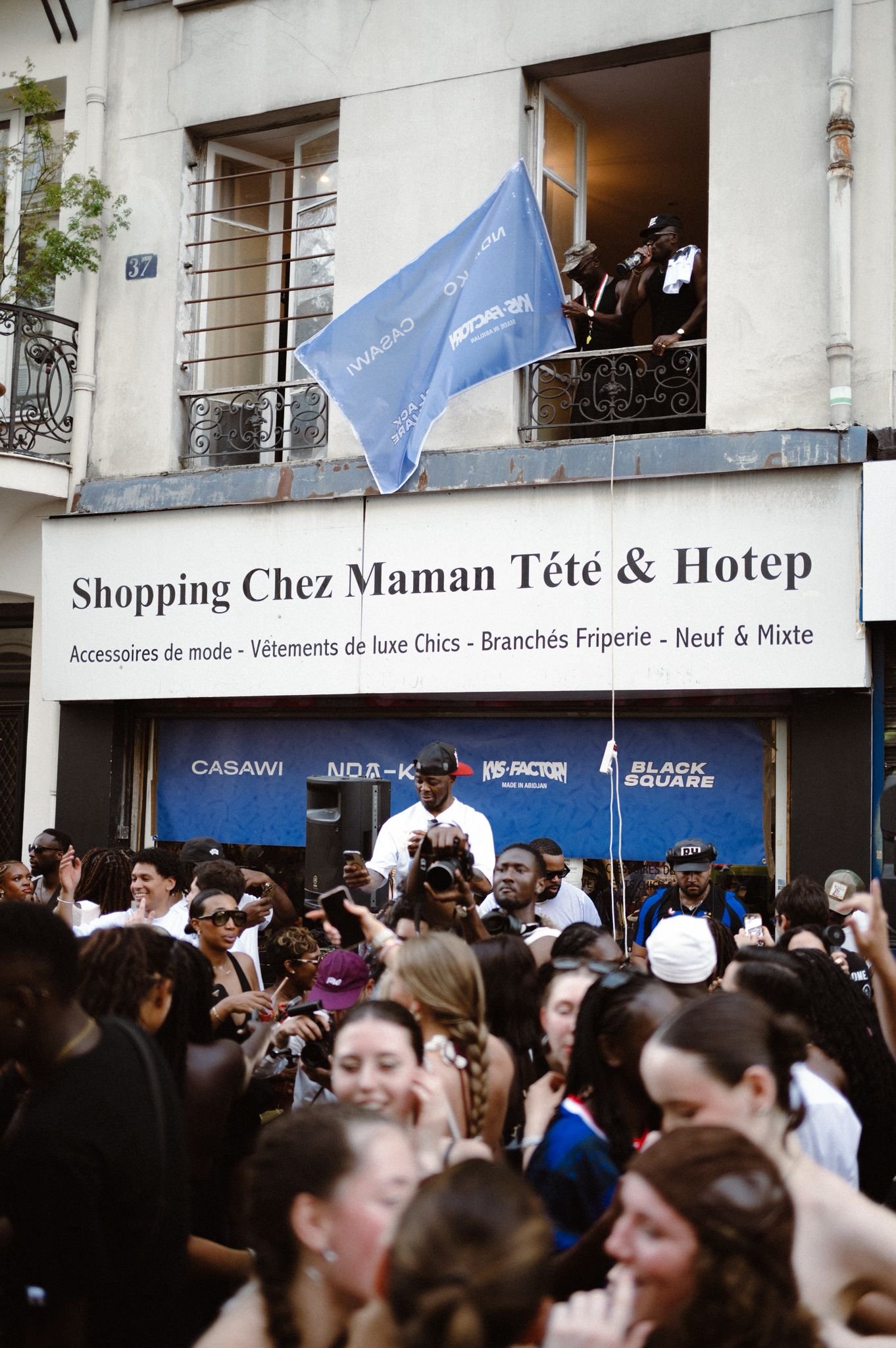 A crowd of people gathered outside a building with a sign that reads "Shopping Chez Maman Tété & Hotep". Inside the building, two men are standing on a balcony, one of whom is drinking from a bottle and another holding a blue flag with white text.