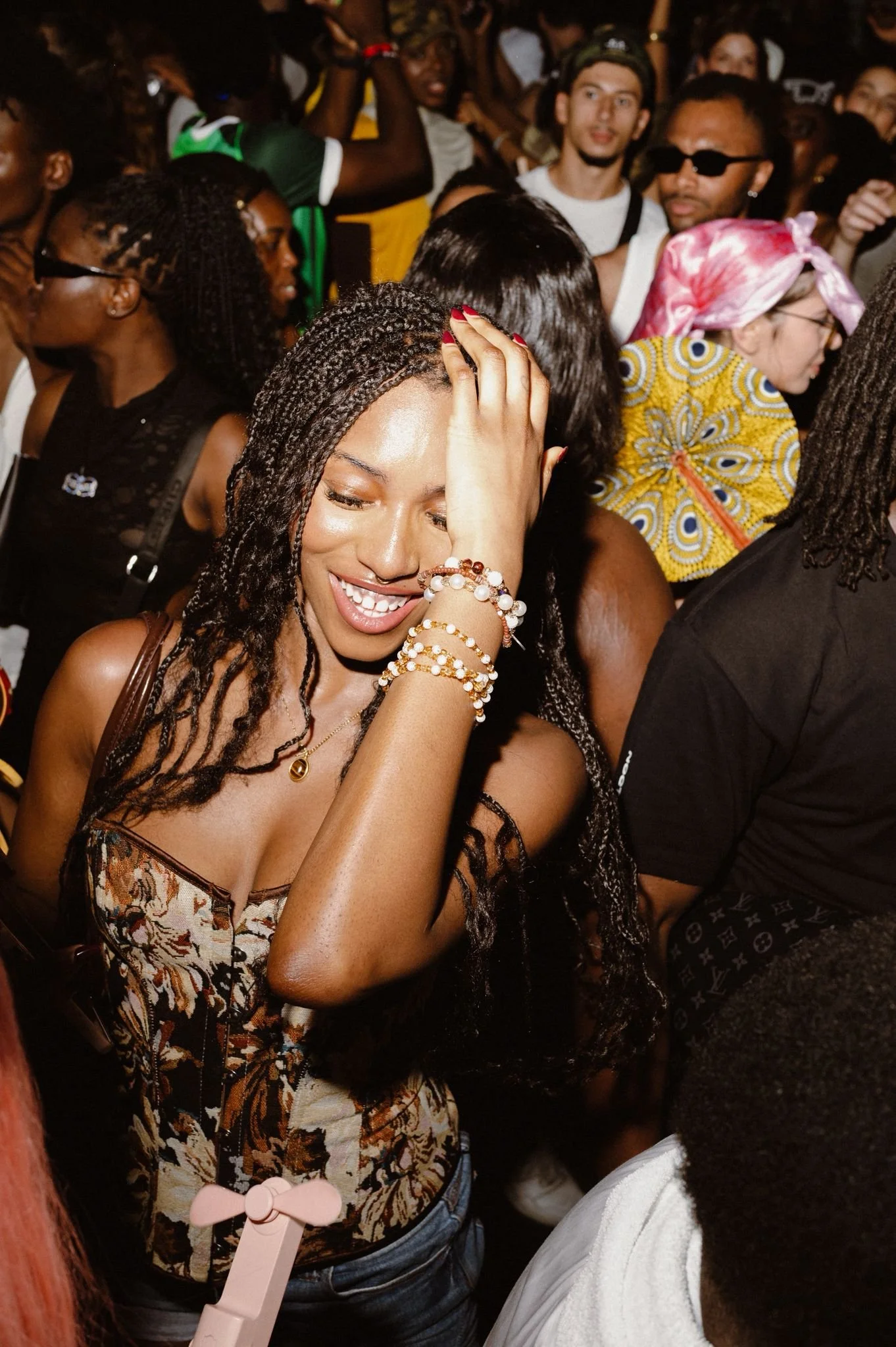 A crowded dance floor with diverse individuals, including a woman with long braided hair, wearing a floral top and multiple bracelets, smiling and touching her face, surrounded by others at a lively social event.
