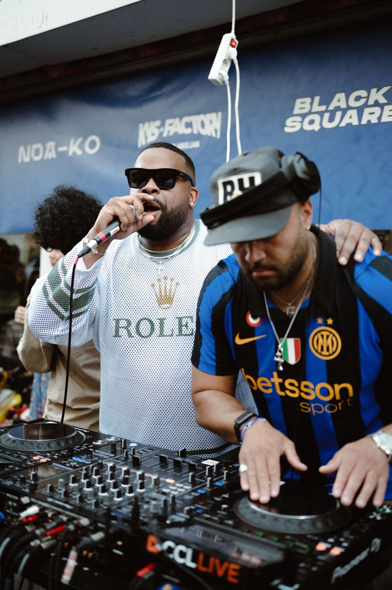 Two men DJing at a party, one wearing a white Rolex shirt and sunglasses, the other in a black and blue soccer jersey, with DJ equipment in front of them.