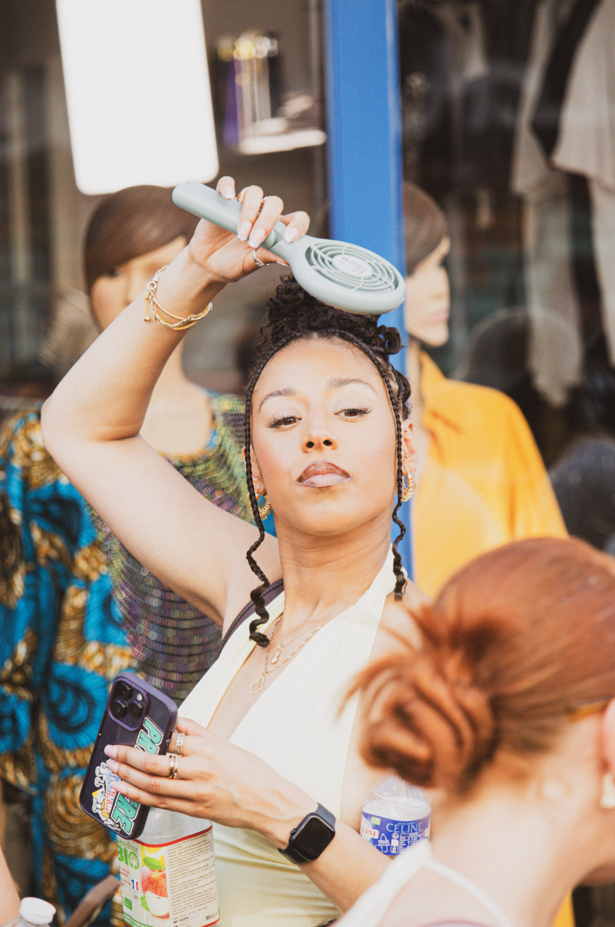 A woman with braided hair holds a green handheld fan above her head while holding a smartphone in her other hand. She is wearing a white top and jewelry, and is surrounded by people and mannequins in a store window display.
