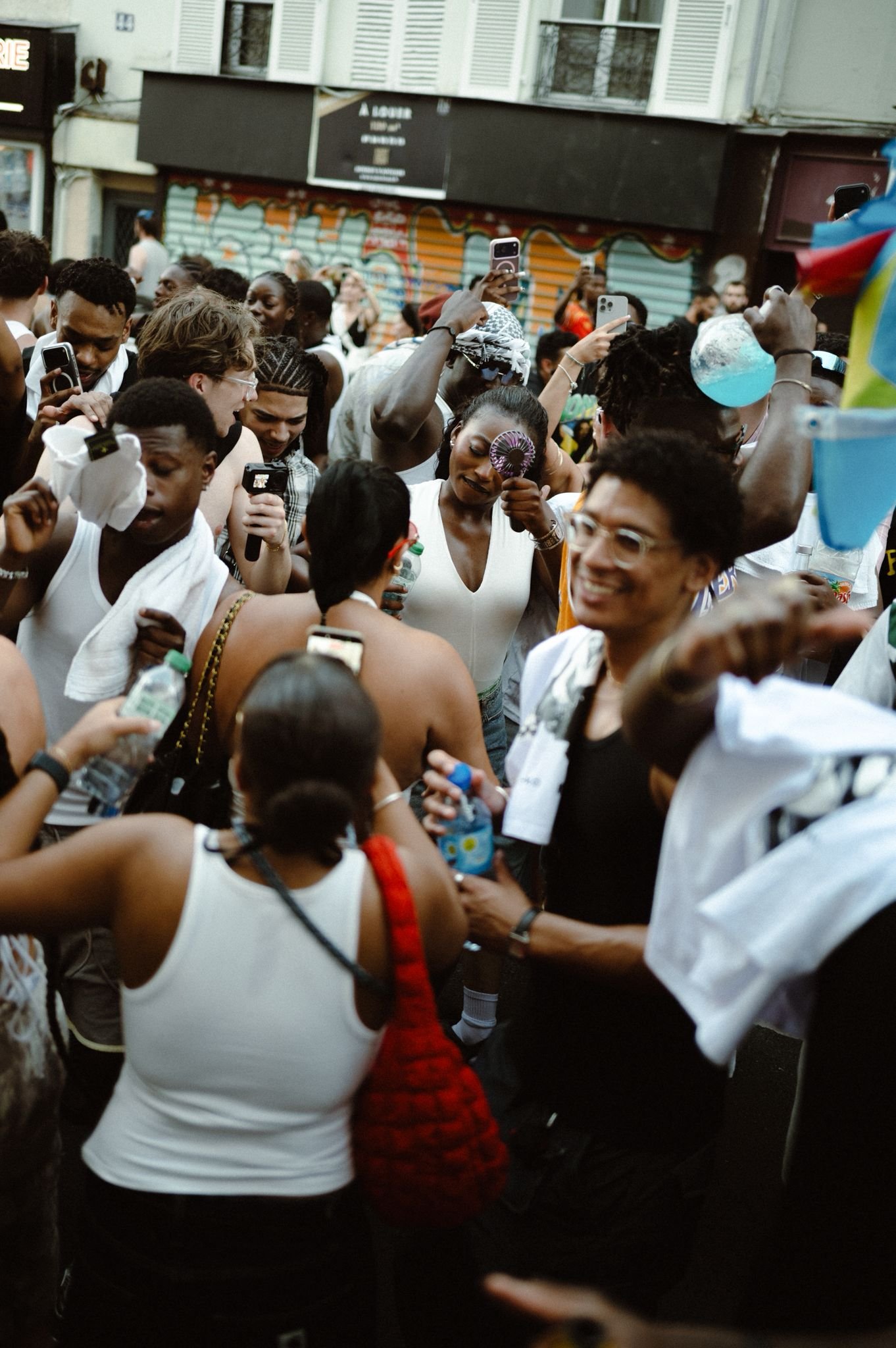 A crowd of people gathered outdoors on a street, many holding up phones and water bottles, with some wearing white tank tops. The background features a graffiti-covered wall and storefronts.