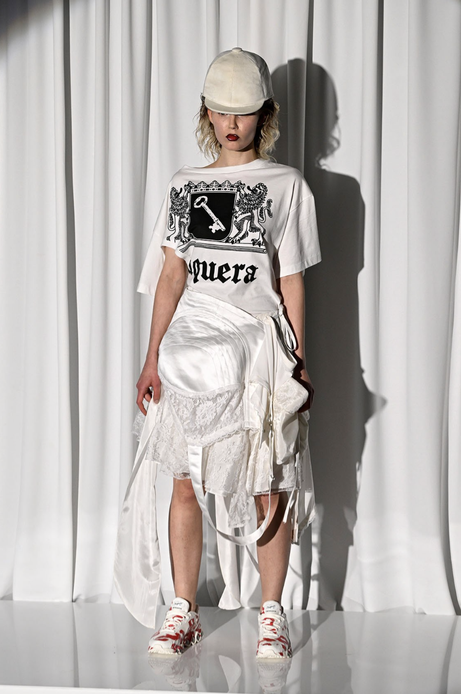 A young woman with curly blonde hair wearing a beige cap, a white T-shirt with a black crest and the text 'muerA', a large white satin skirt with lace and ribbon details, and white sneakers with red accents, standing in front of white curtains.