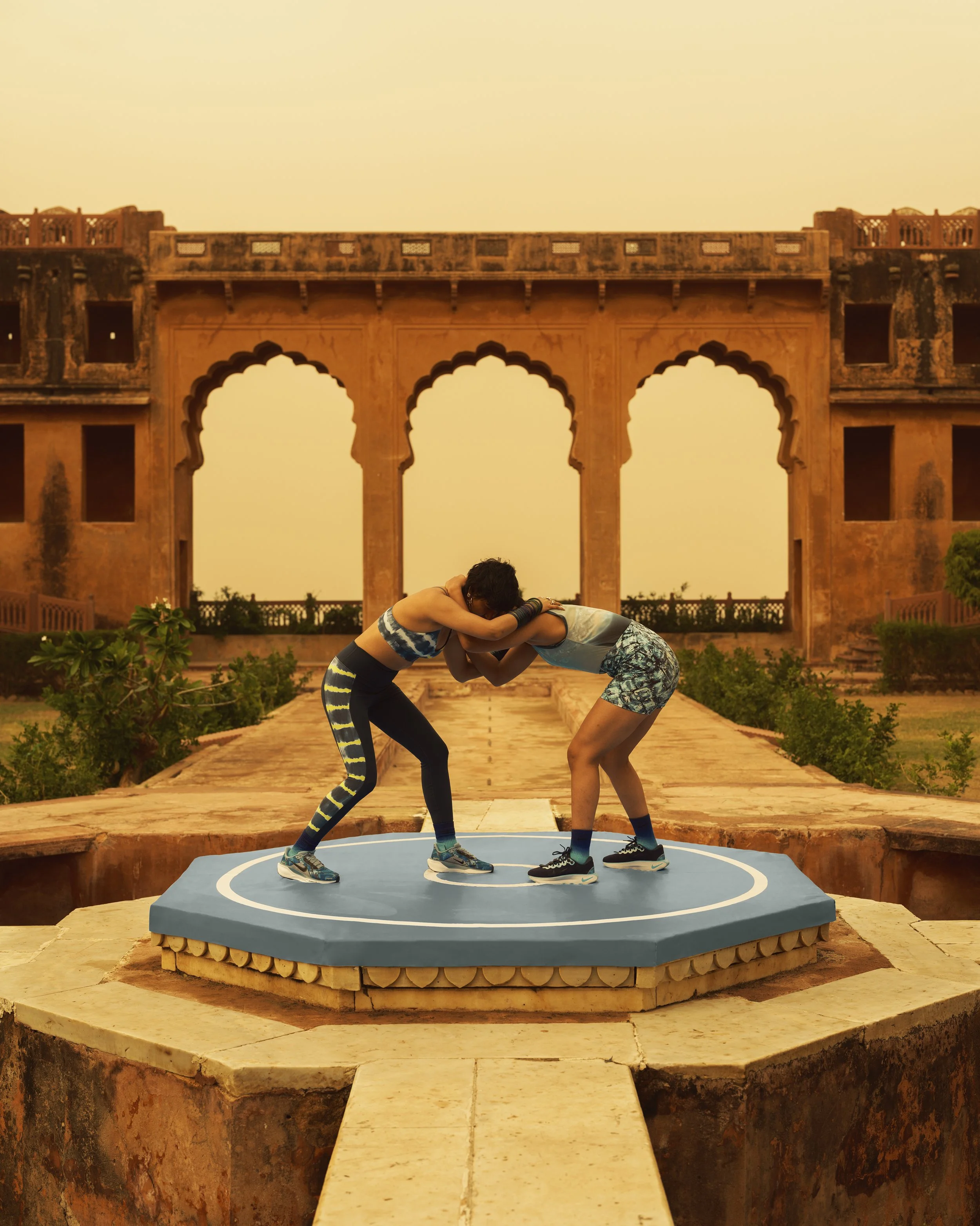 Two women practicing wrestling or a combat sport on a raised platform outdoors, with an ancient, ornate building in the background during sunset.