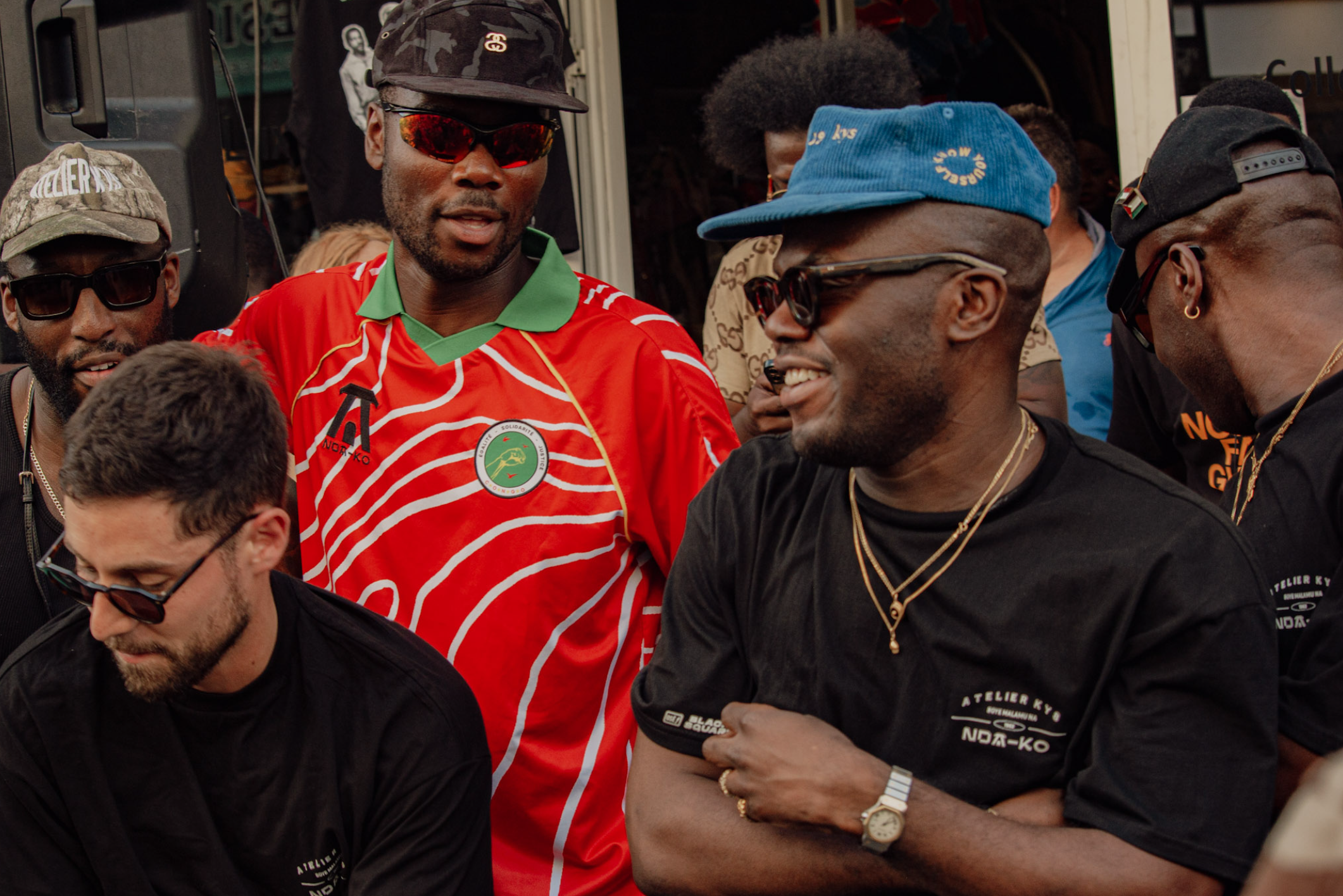 Group of men gathered together, wearing sunglasses, caps, and casual clothing, in an outdoor setting.