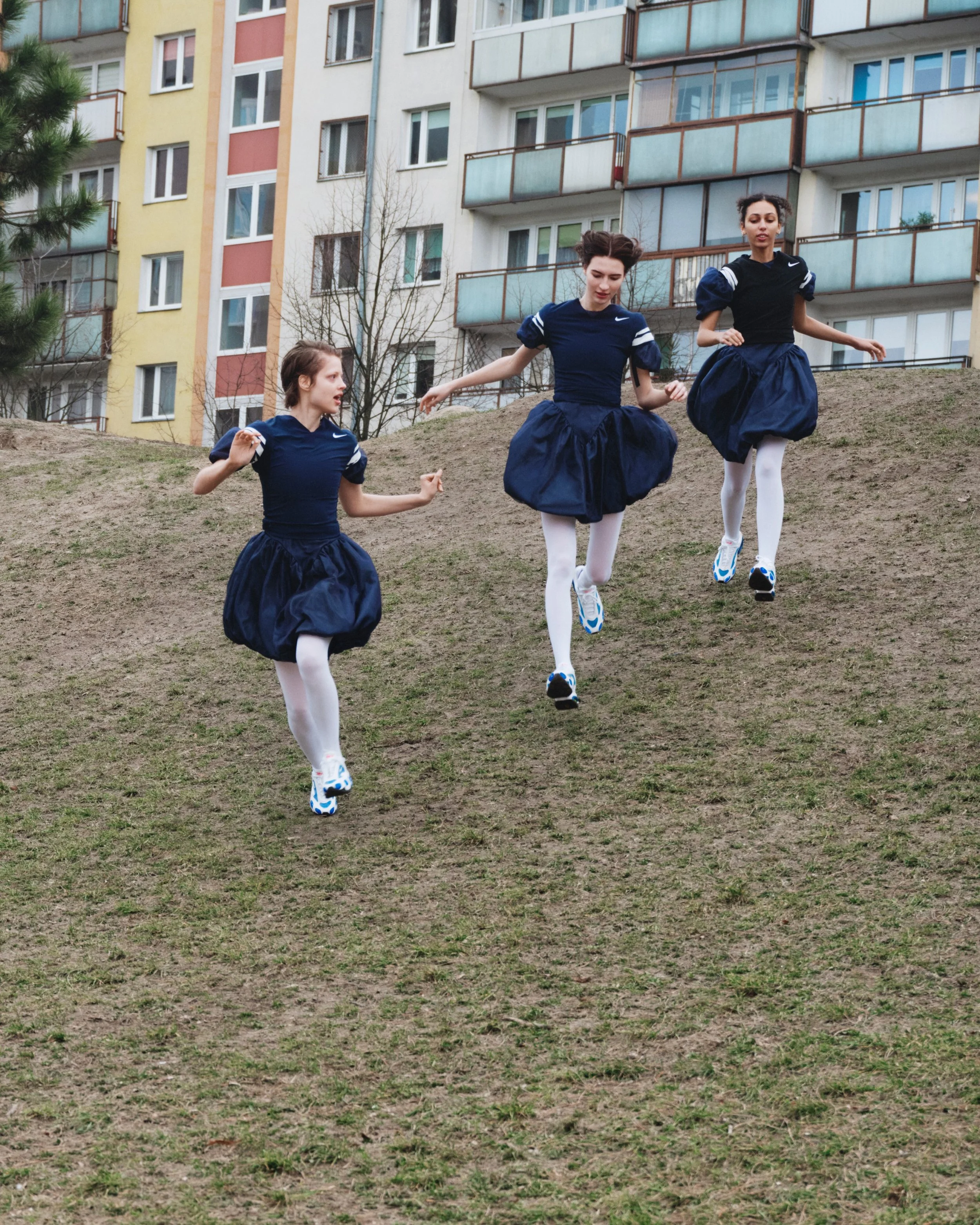 Three young women dressed in dark blue dance costumes with puffy skirts, white tights, and running shoes, practicing dance on a grassy hill in front of an apartment building.