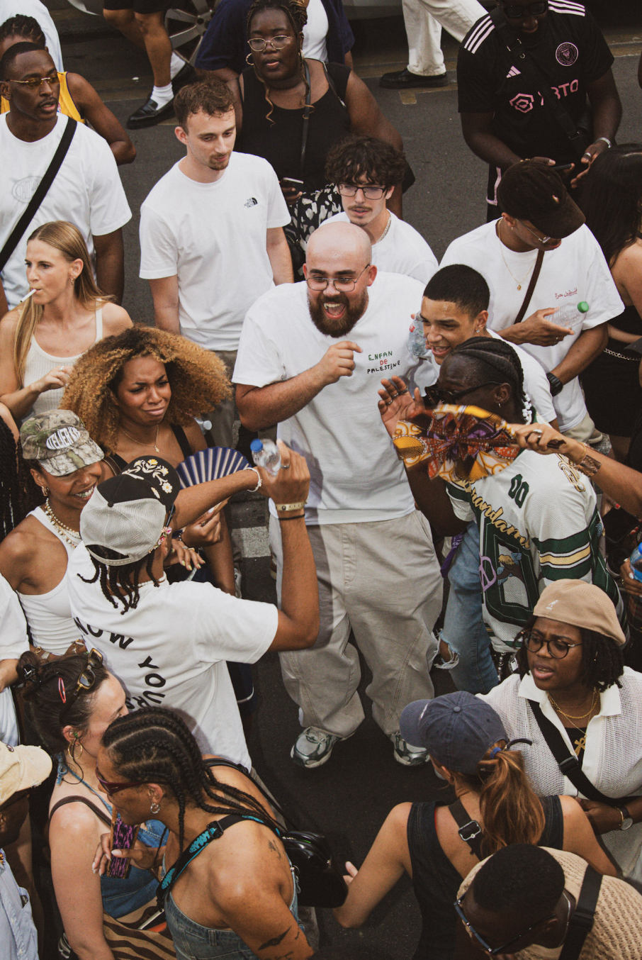 A diverse group of people gathered in a street, engaging in conversations and smiling, with some holding water bottles and others wearing casual summer clothing.