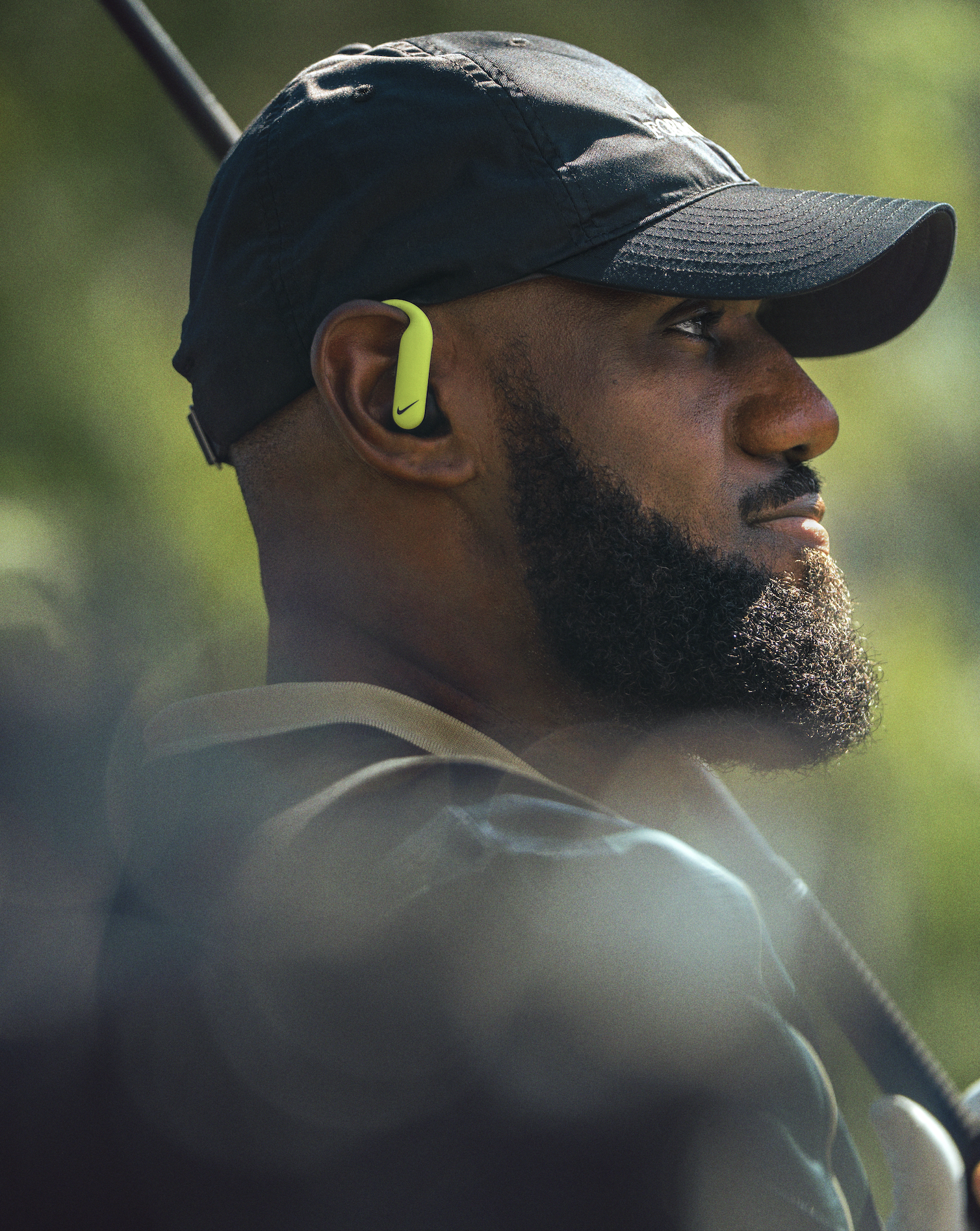 A close-up side profile of a man wearing a black cap and a yellow Nike wireless earbud, with a golf club behind him, likely outdoors on a sunny day.