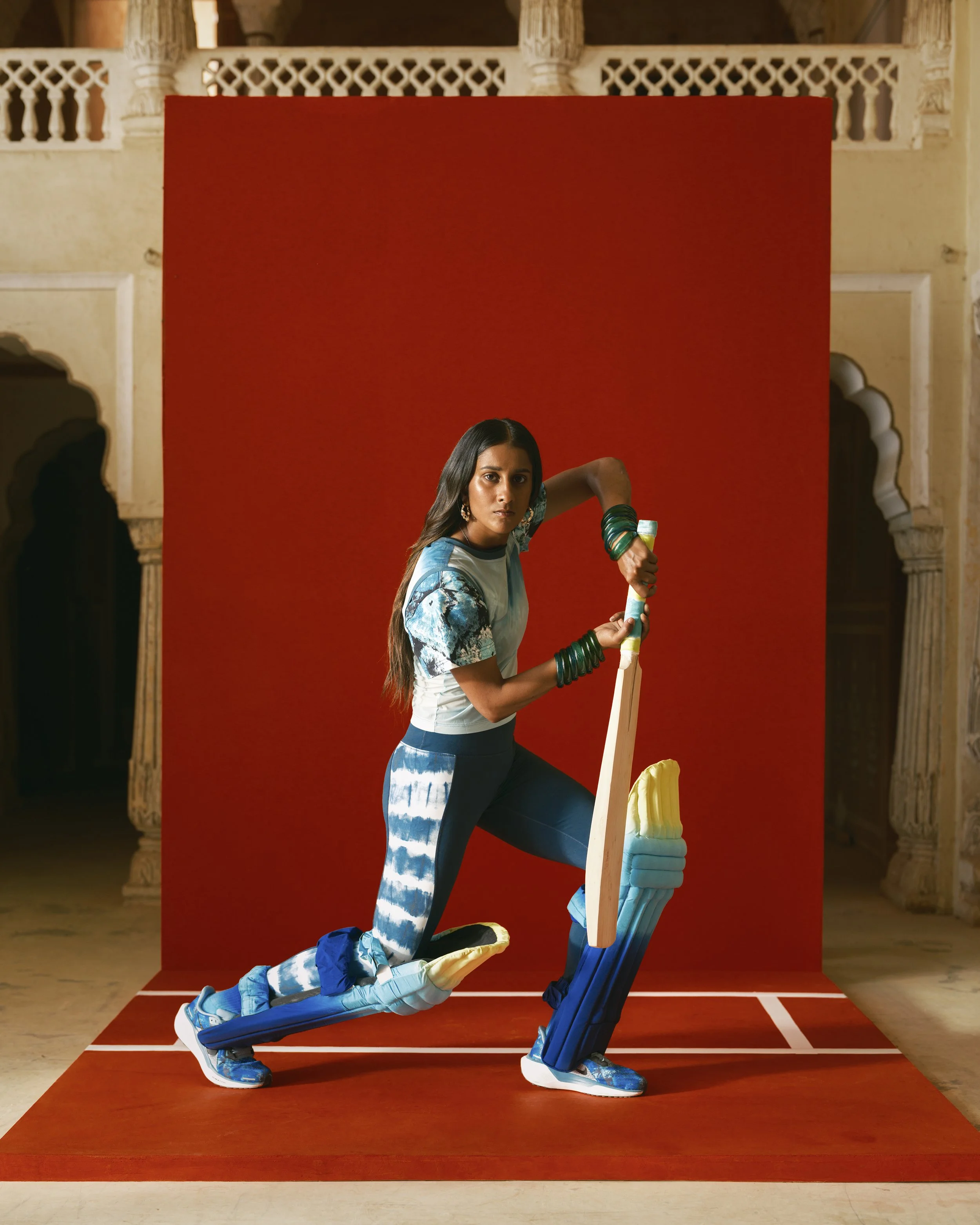 A woman in blue and white athletic clothes and green bangles poses on a red mat in front of a red backdrop, with traditional architectural details in the background.
