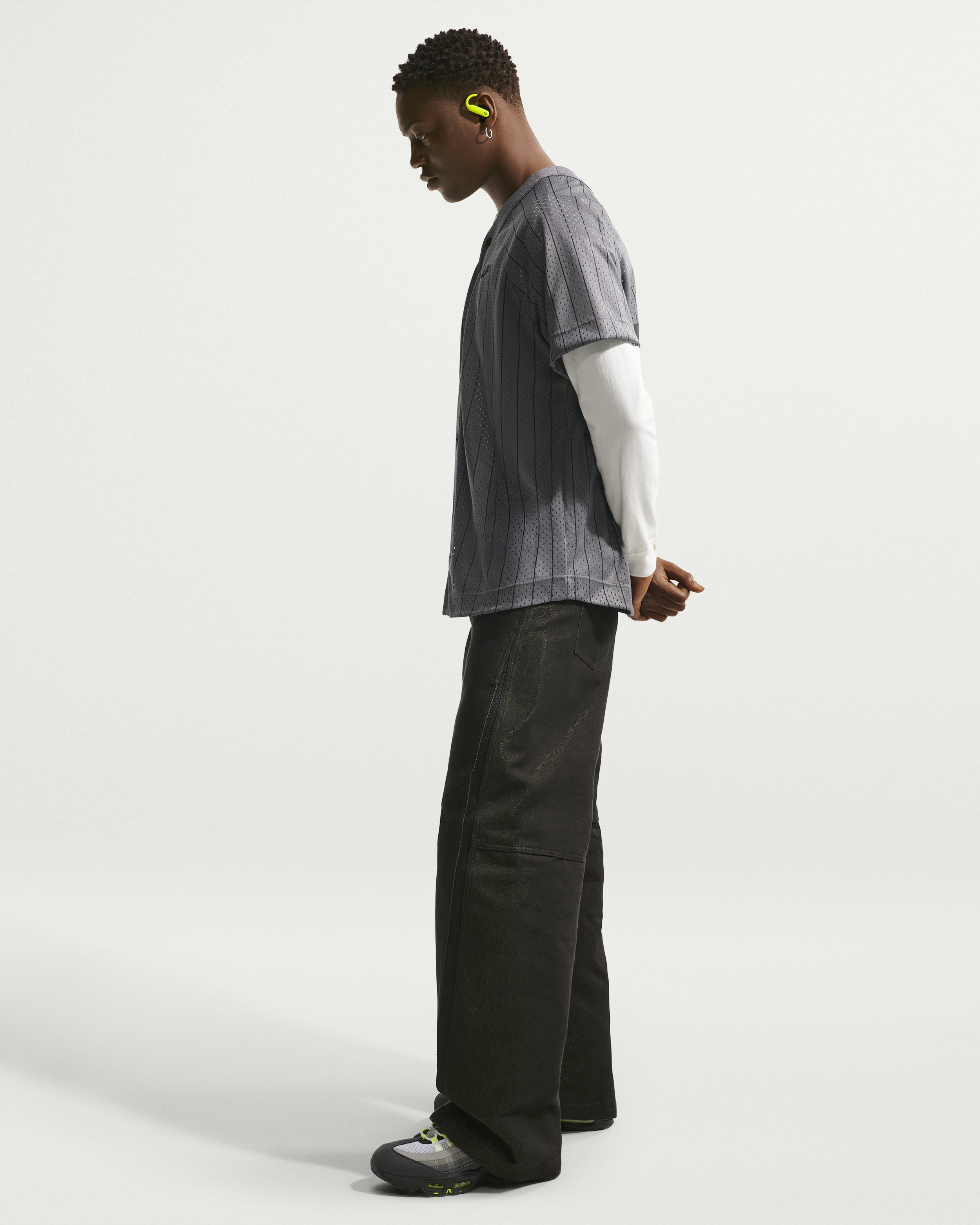 Side view of a person wearing a grey patterned shirt over a white long sleeve shirt, black pants, and black sneakers with grey and neon yellow accents, standing against a plain white background. They are wearing yellow wireless earbuds and have their hands clasped behind their back while looking down.