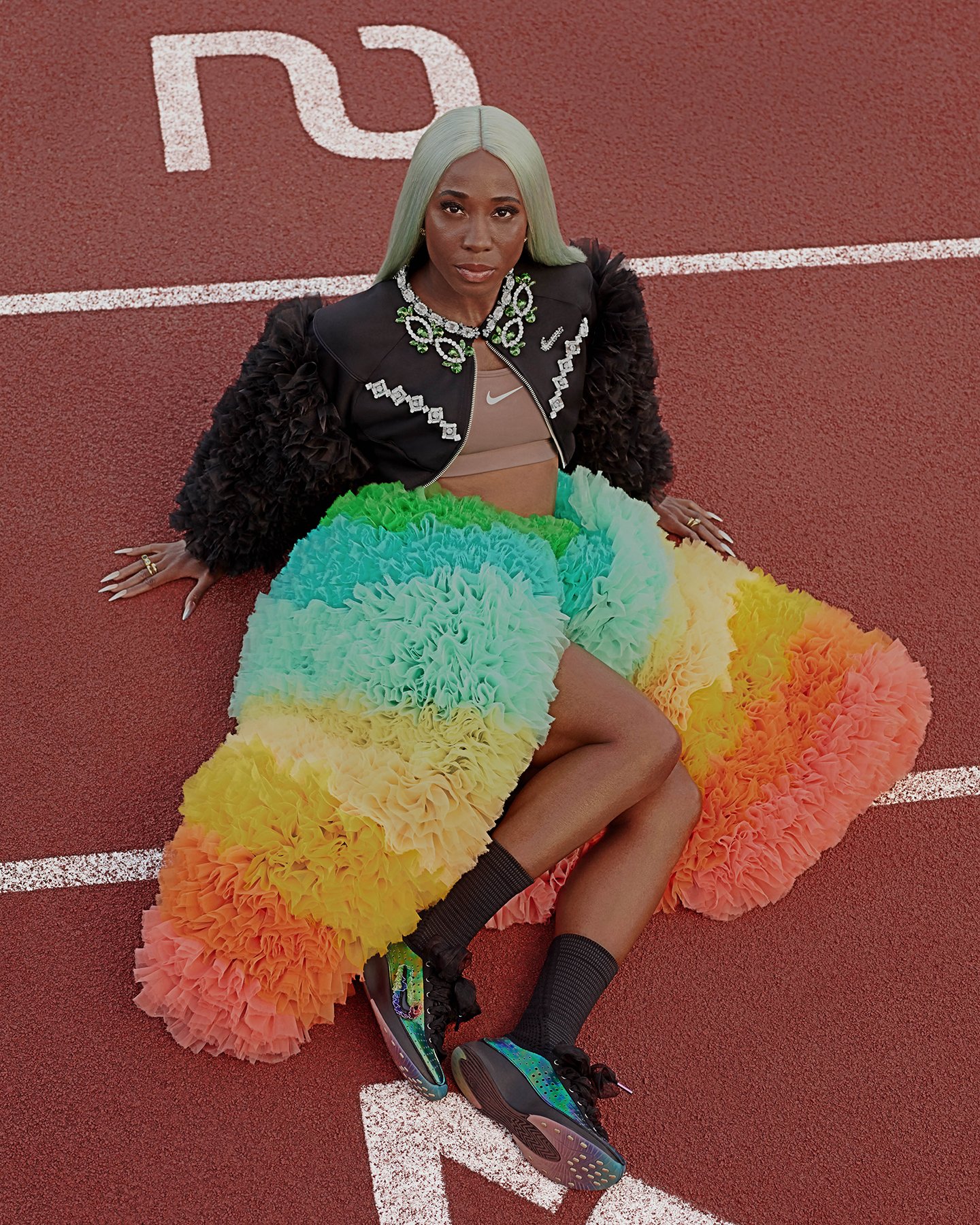 A woman with long pastel green hair sitting on a red track field, wearing a black jacket with silver embellishments, a beige Nike sports bra, and a colorful ruffled skirt in shades of blue, green, yellow, orange, and pink, with black high-top sneakers.