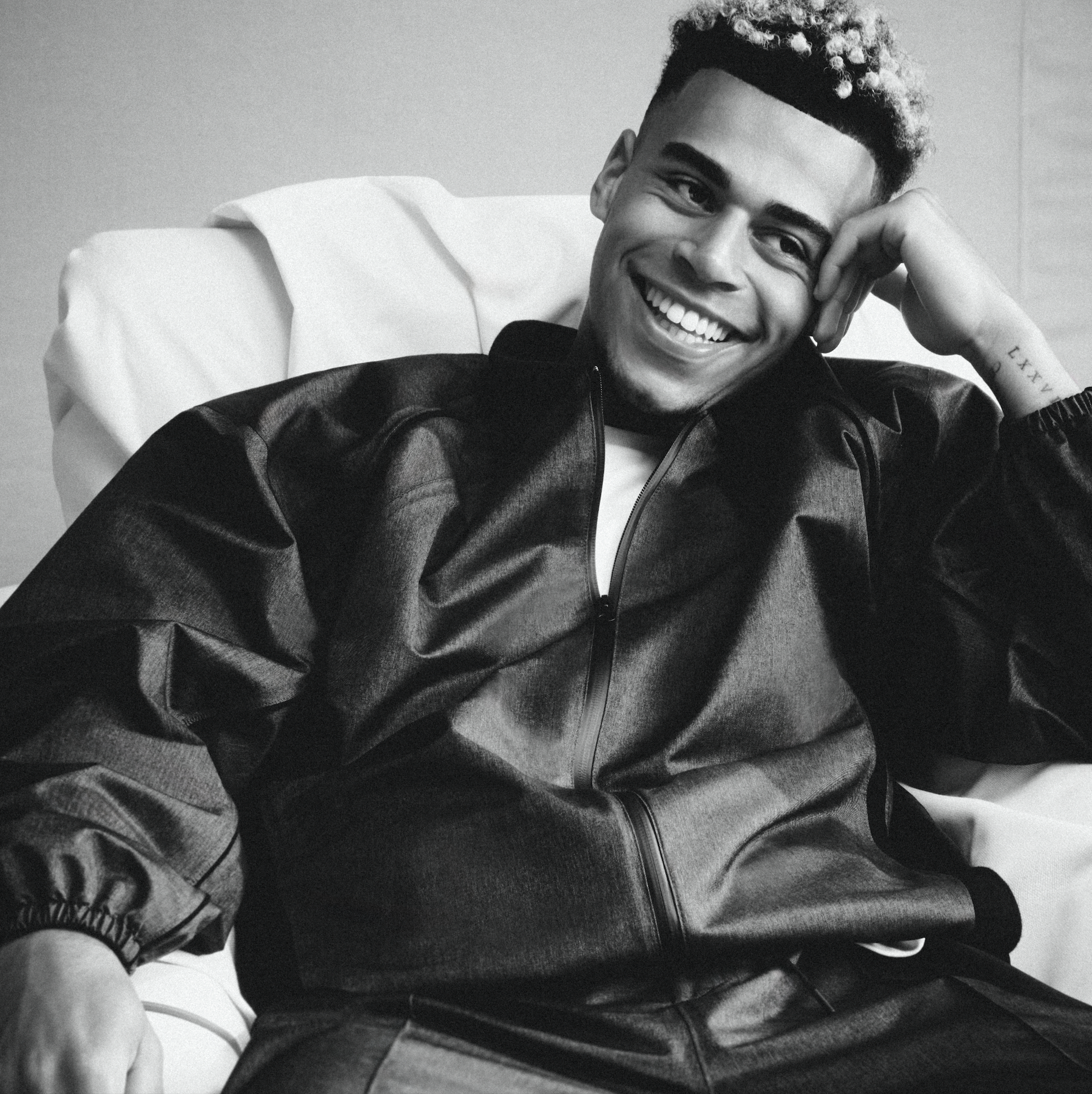 A smiling young man with curly hair, a headband, and tattoos, sitting casually in a relaxed posture.