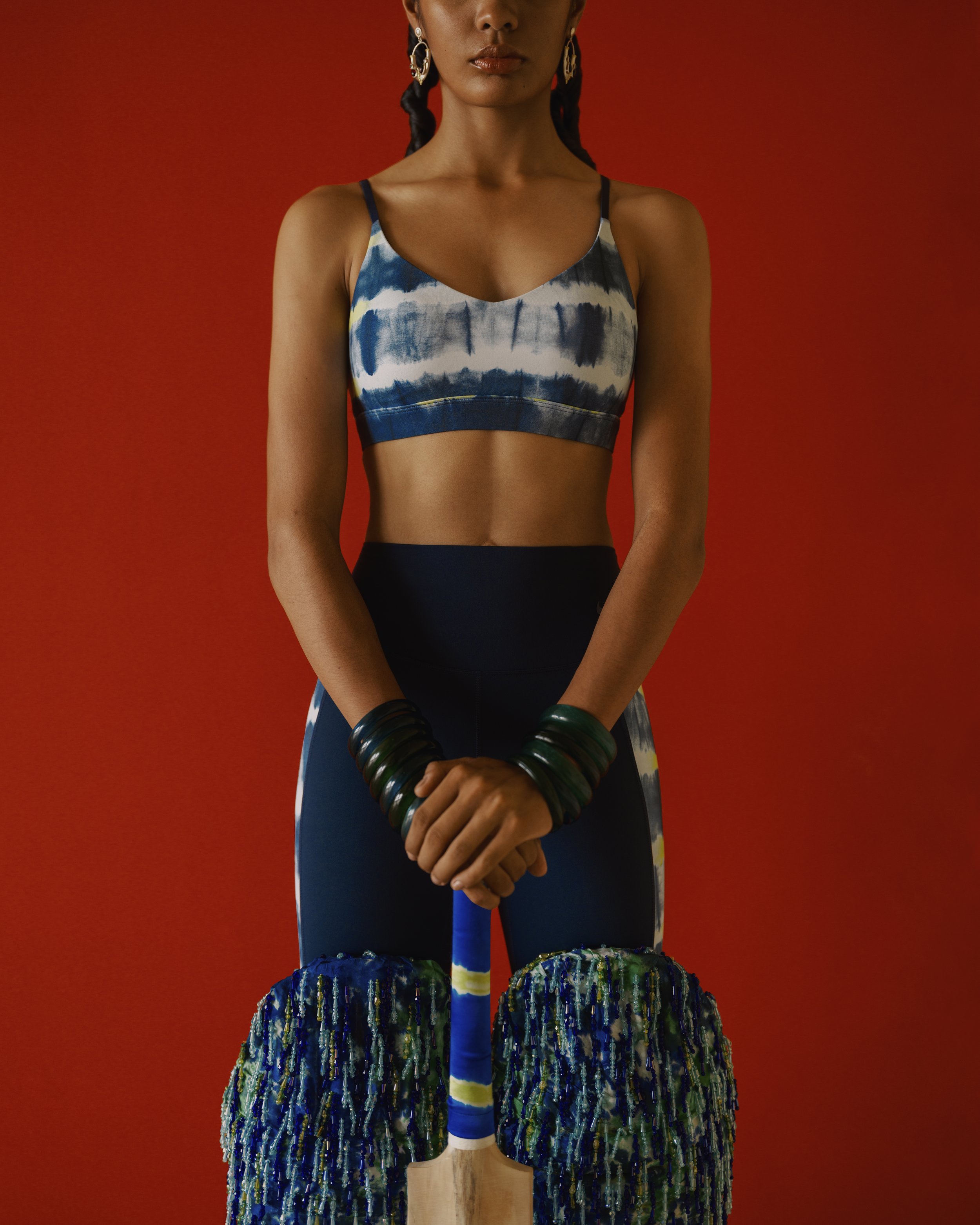 A person standing against a red background, wearing a blue and white tie-dye sports bra and matching leggings, with jewelry and black wristbands.