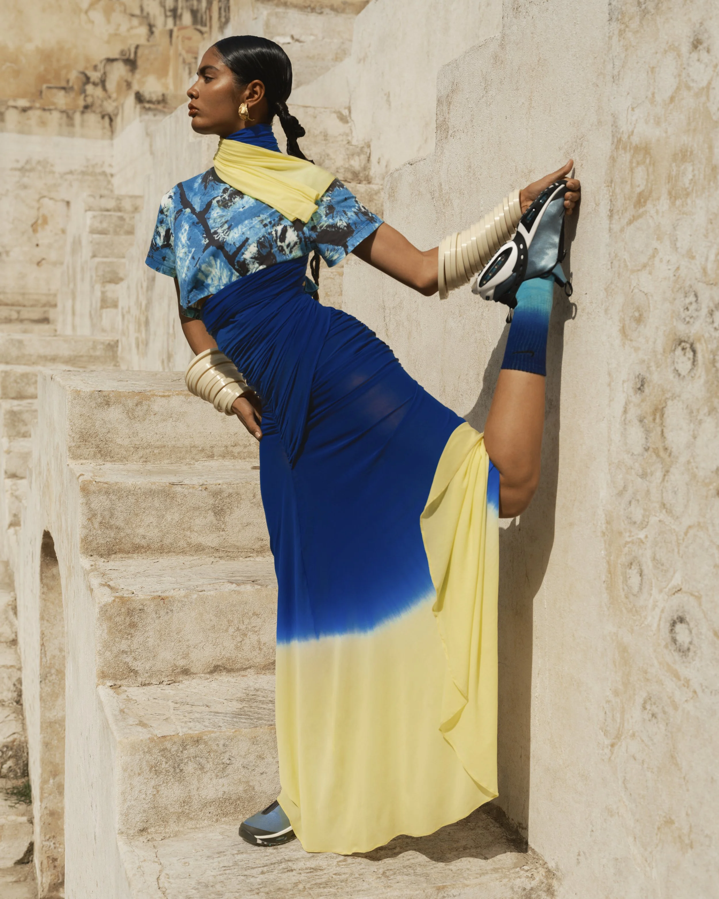 A woman sitting on stone steps against a beige stone wall, wearing a blue, yellow, and black outfit with modern sneakers, with a calm and focused expression.