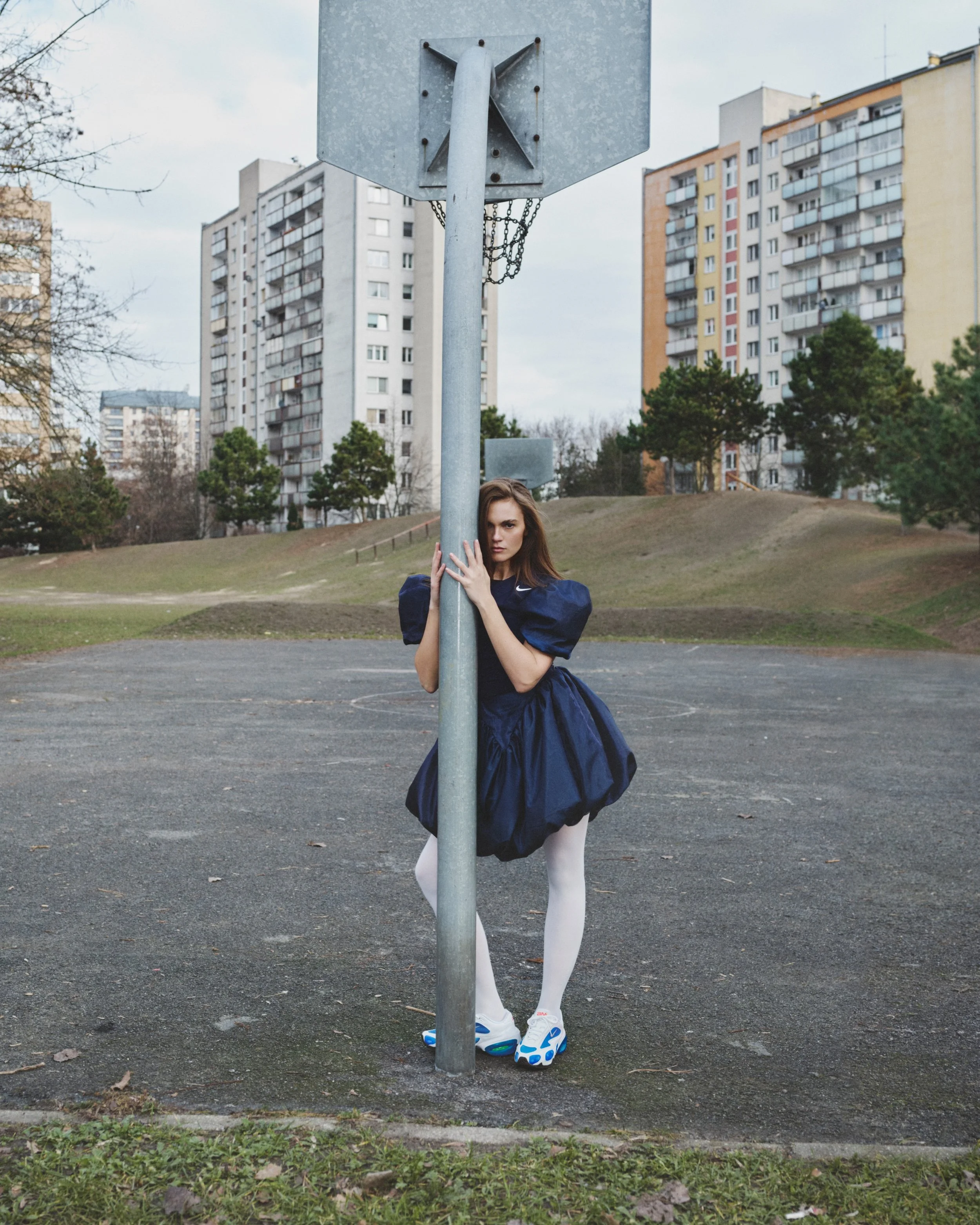 A woman dressed in a dark blue vintage-style dress with puffed sleeves, white tights, and colorful athletic shoes, stands behind an basketball hoop on an outdoor court, holding the pole that supports the backboard and hoop, with high-rise apartment buildings in the background.