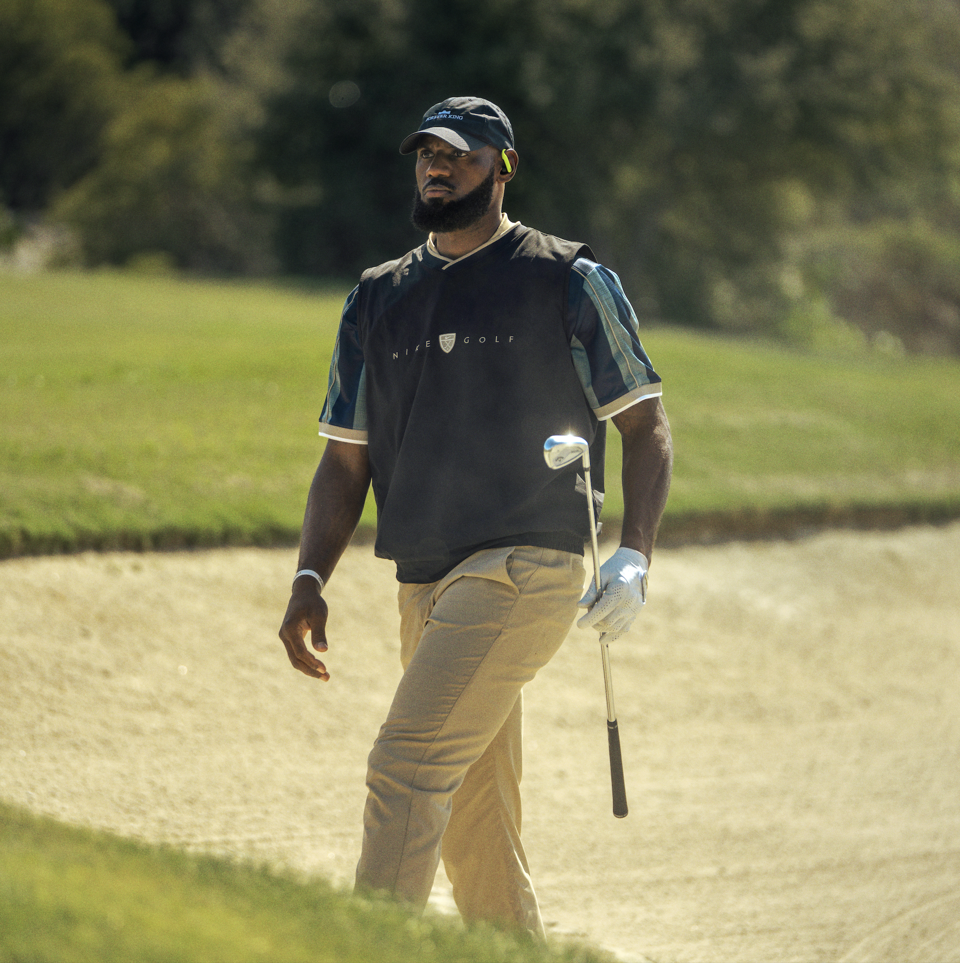 Man walking on a golf course holding a golf club, wearing a cap, plaid shirt, gloves, and wireless earbud, with trees blurred in the background.