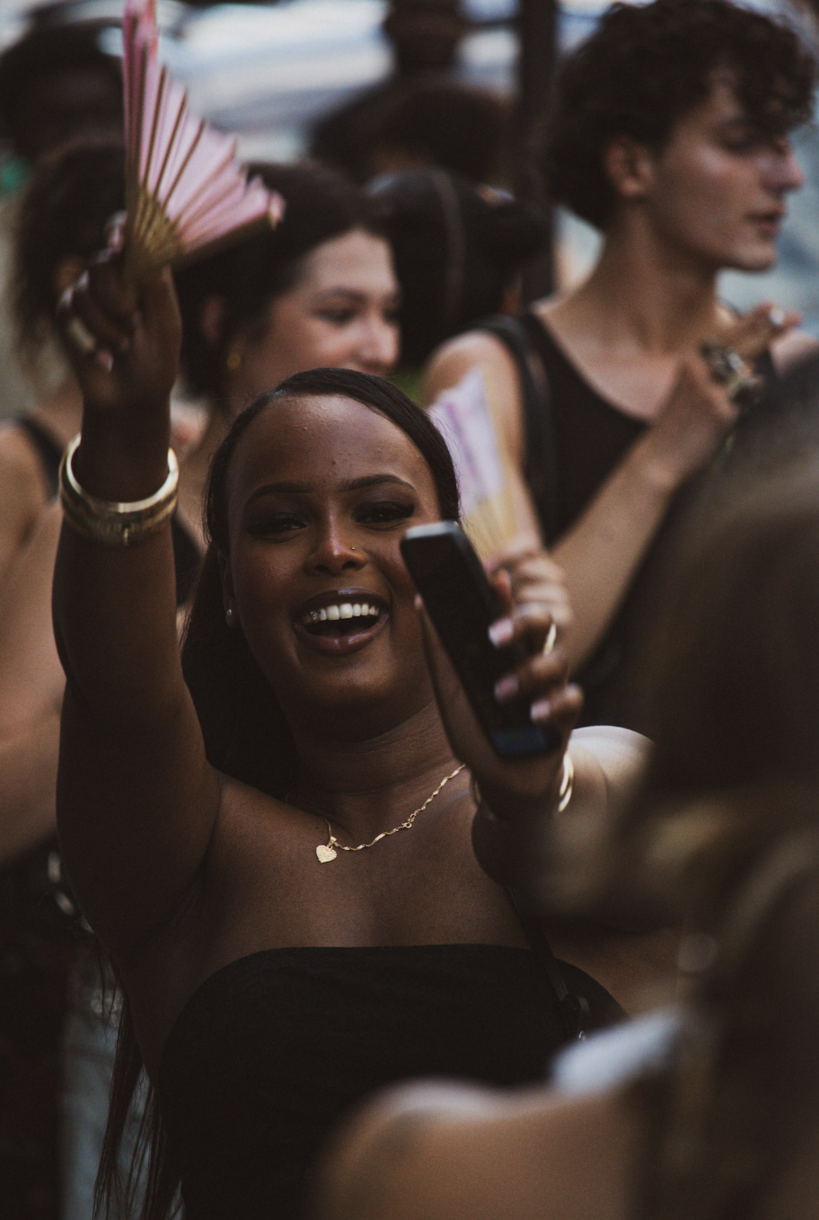 A woman with dark hair smiling and taking a selfie at a lively outdoor event with other people around.