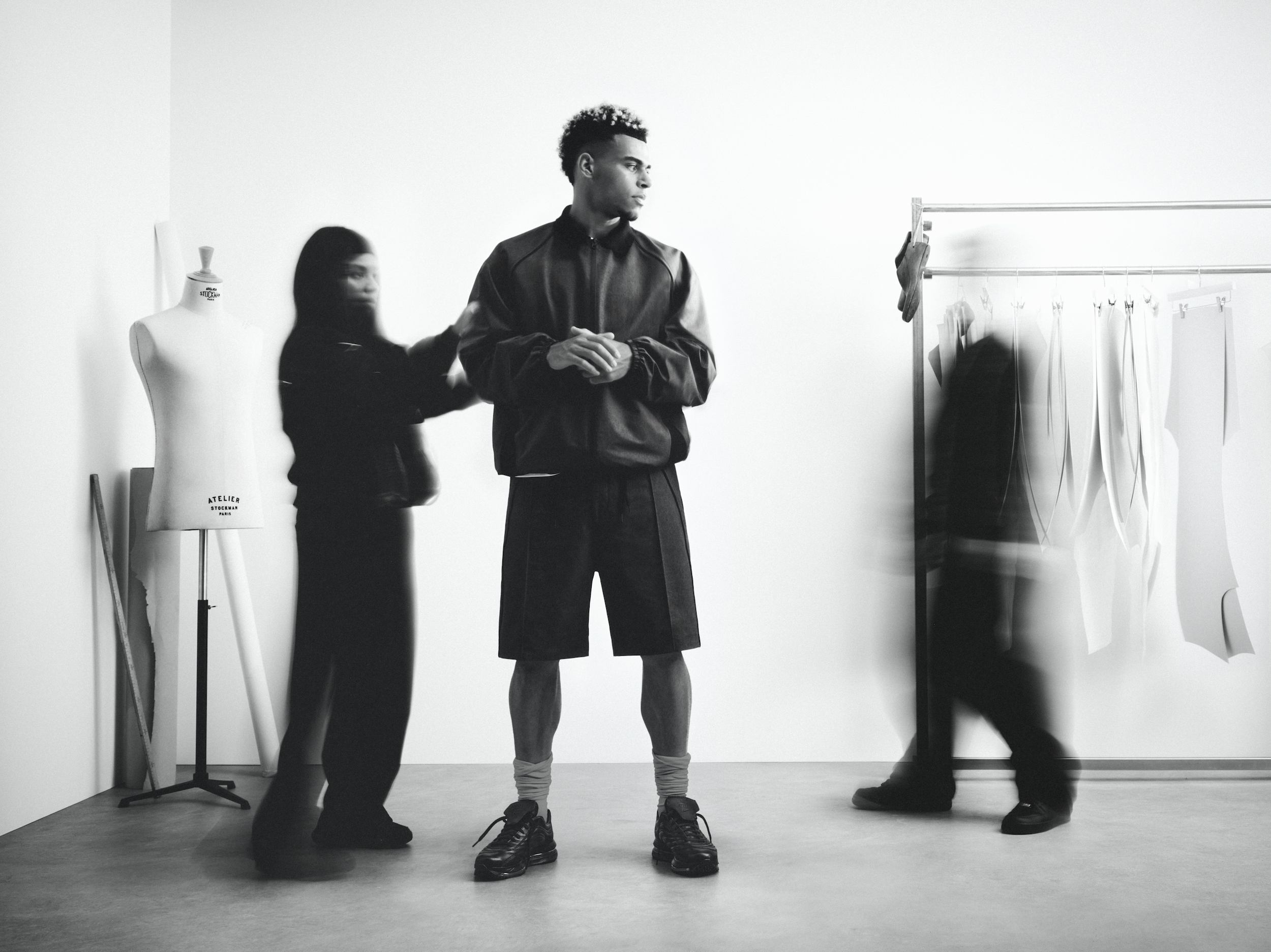 A black and white photo of a young man standing in a studio, with two women near him, and clothing racks on the right side. One woman is adjusting the young man's jacket, and they are surrounded by a mannequin and clothing racks.