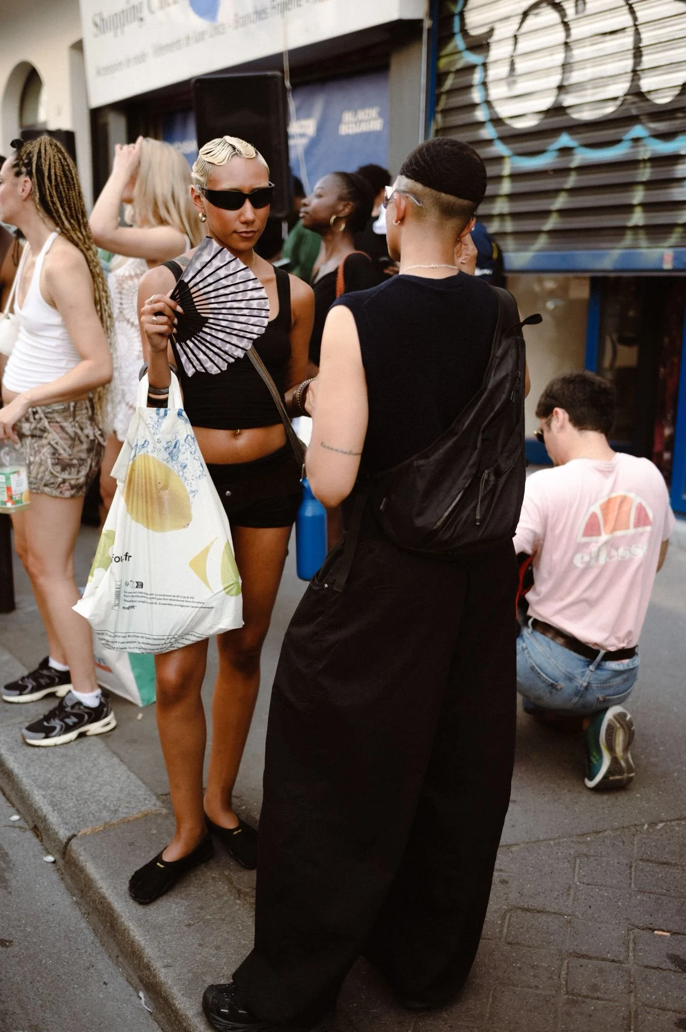 Two women are talking on a city sidewalk, one woman with blonde hair, sunglasses, a black top, and shorts, holding a fan, and the other woman with short hair, glasses, and a black dress, facing each other. Several other people are in the background, some sitting and some standing, near a storefront with a shutter.