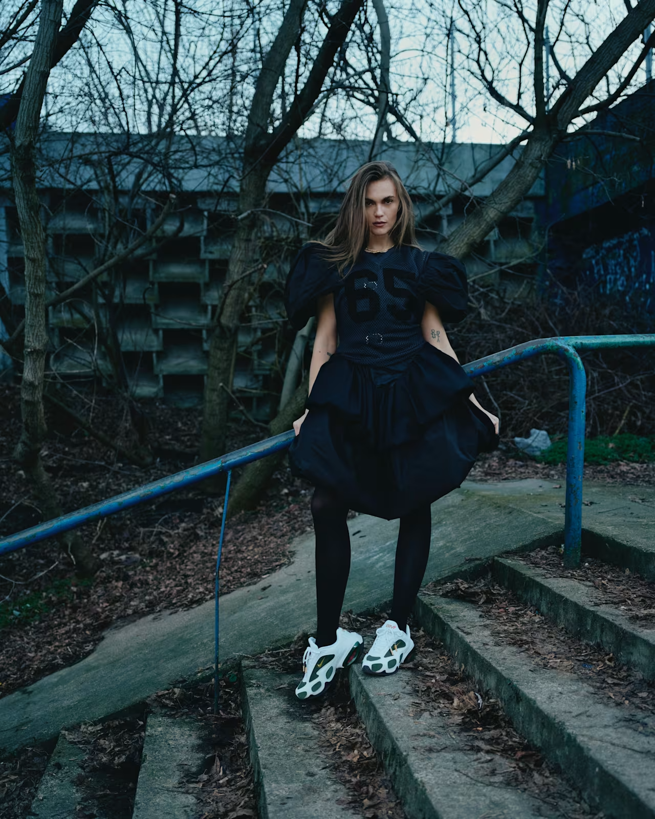 A young woman in a dark sporty outfit and chunky white sneakers stands on outdoor steps near a blue railing, with leafless trees and a building in the background.