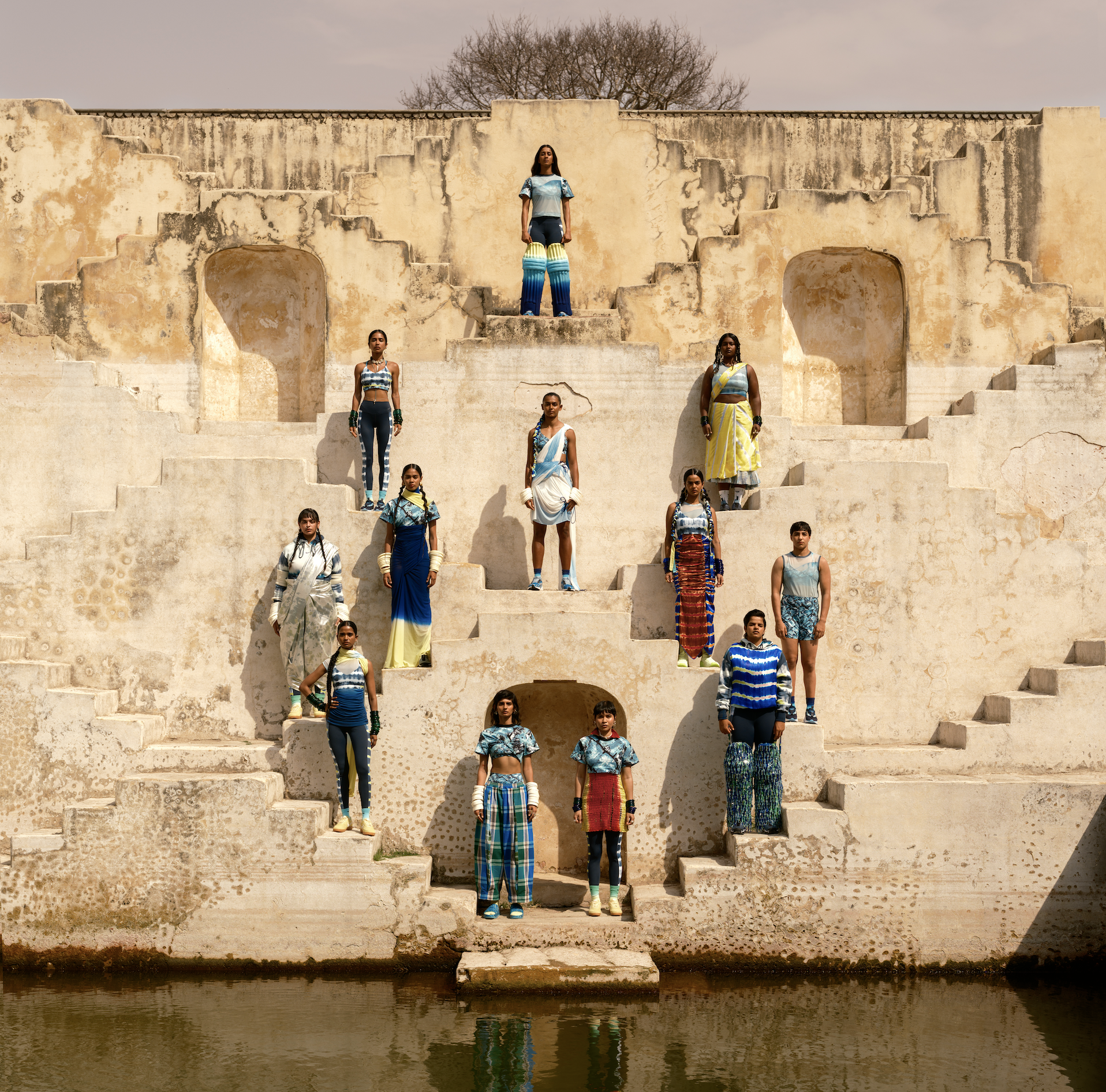 Group of nine people standing on steps of an old, weathered stone structure, with a body of water in the foreground, and a tree in the background. They are wearing colorful, patterned clothing and have various accessories.